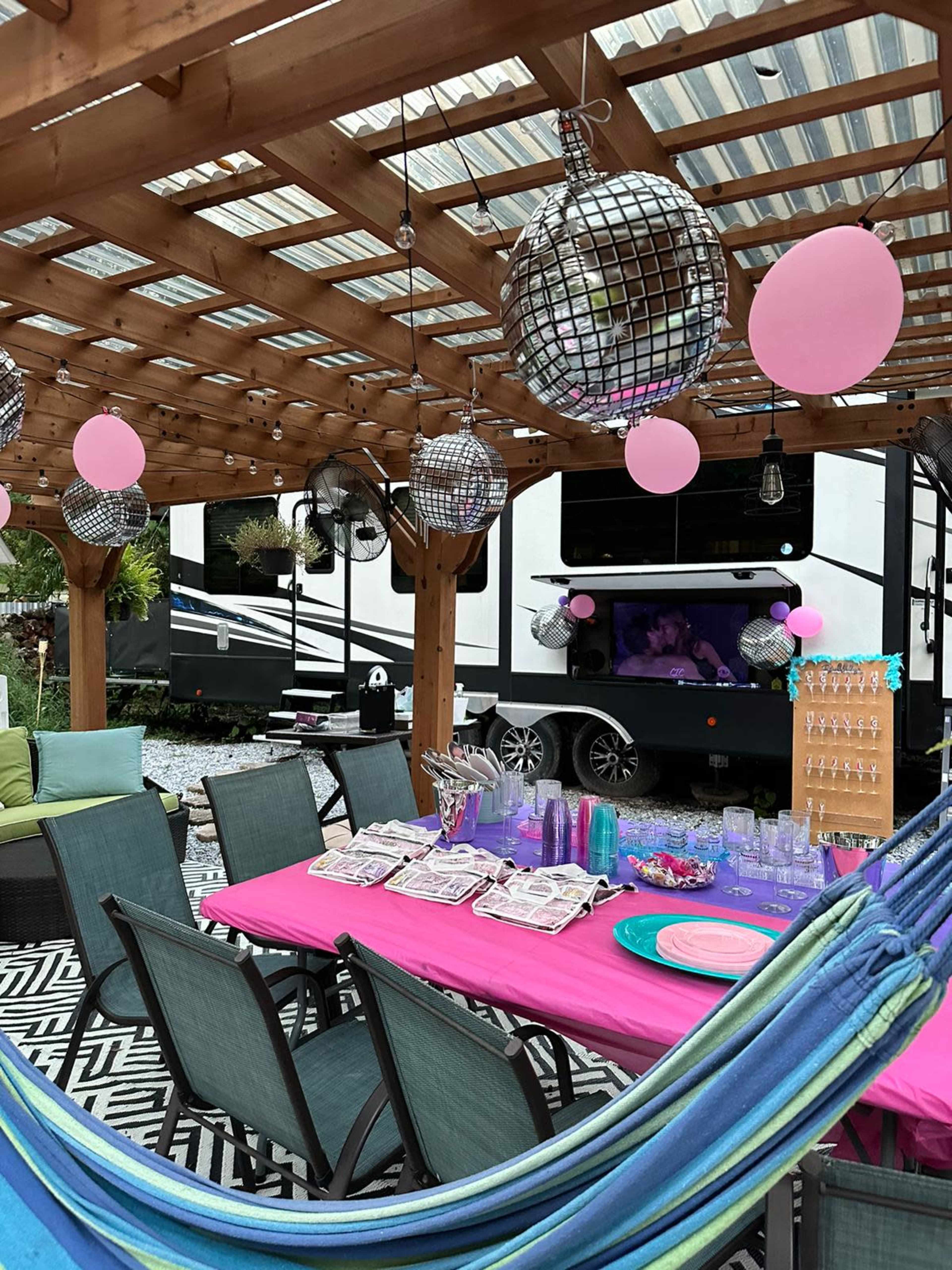 A backyard patio features a colorful table set for a party, adorned with balloons and disco balls, alongside an RV parked in the background.