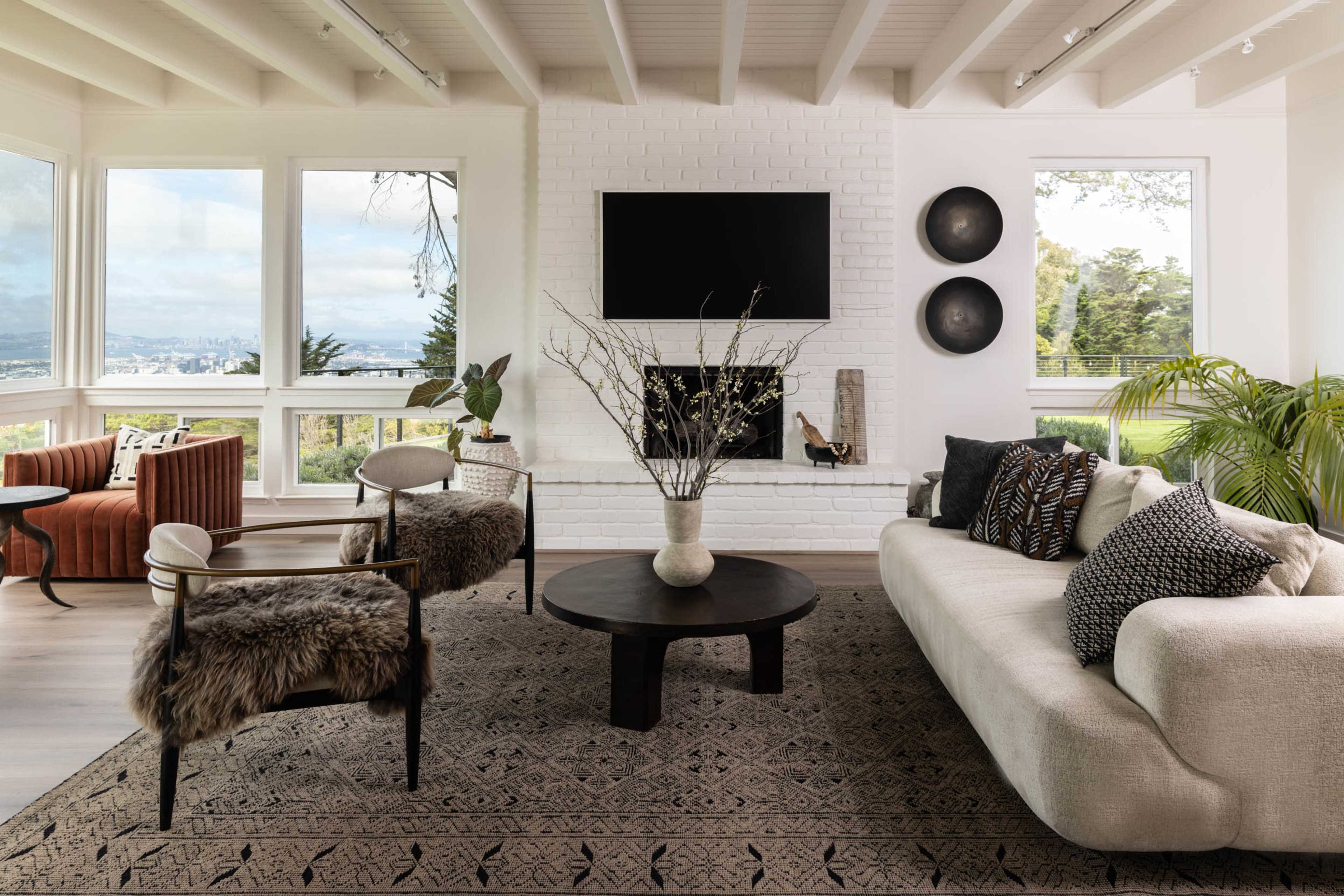 A modern living room features a beige sofa, two accent chairs, a dark round coffee table, and a wall-mounted television above a white-brick fireplace, with large windows showing a scenic view.