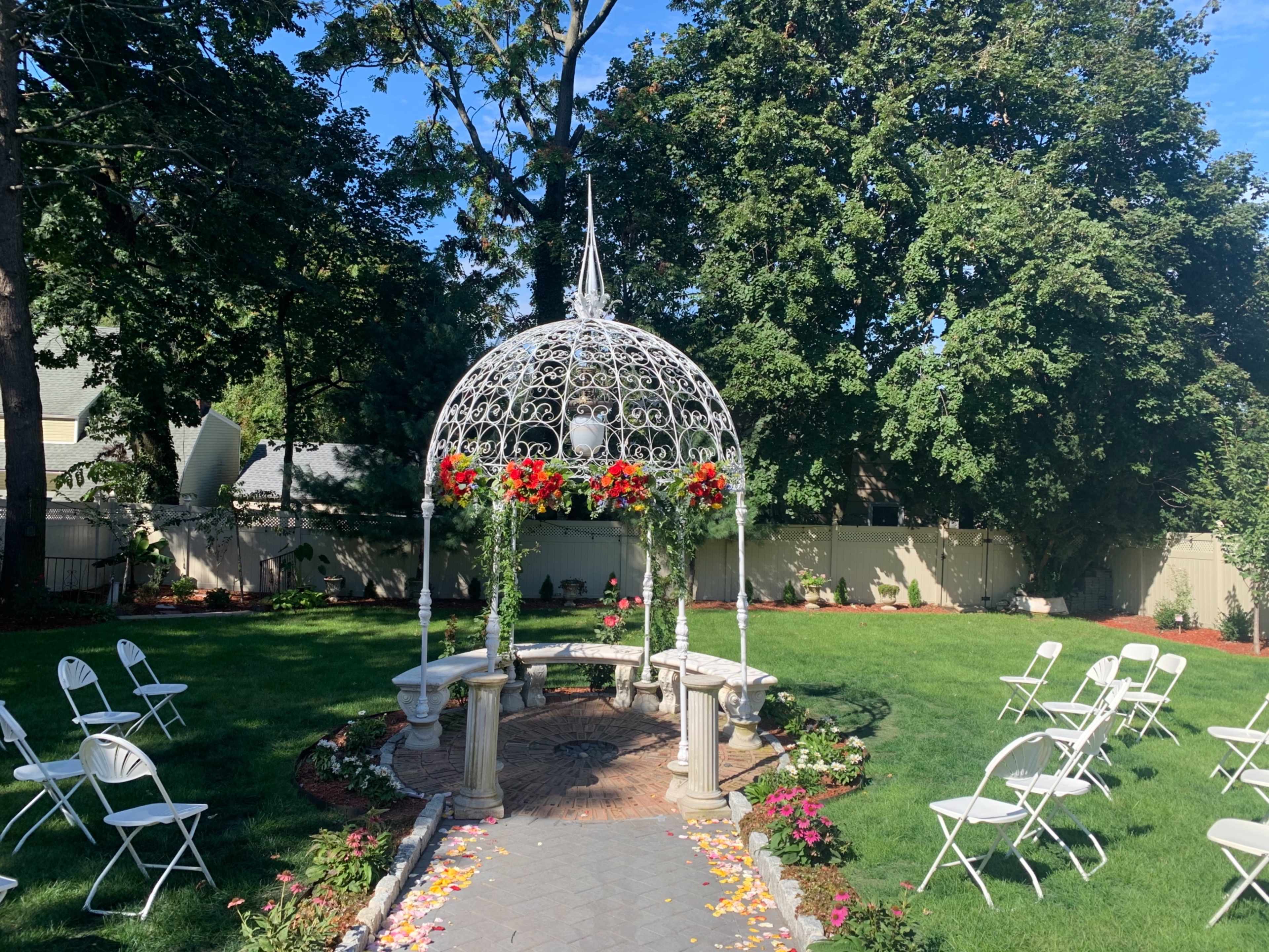 The image shows a landscaped garden with a white gazebo adorned with flowers, surrounded by chairs arranged for an event.