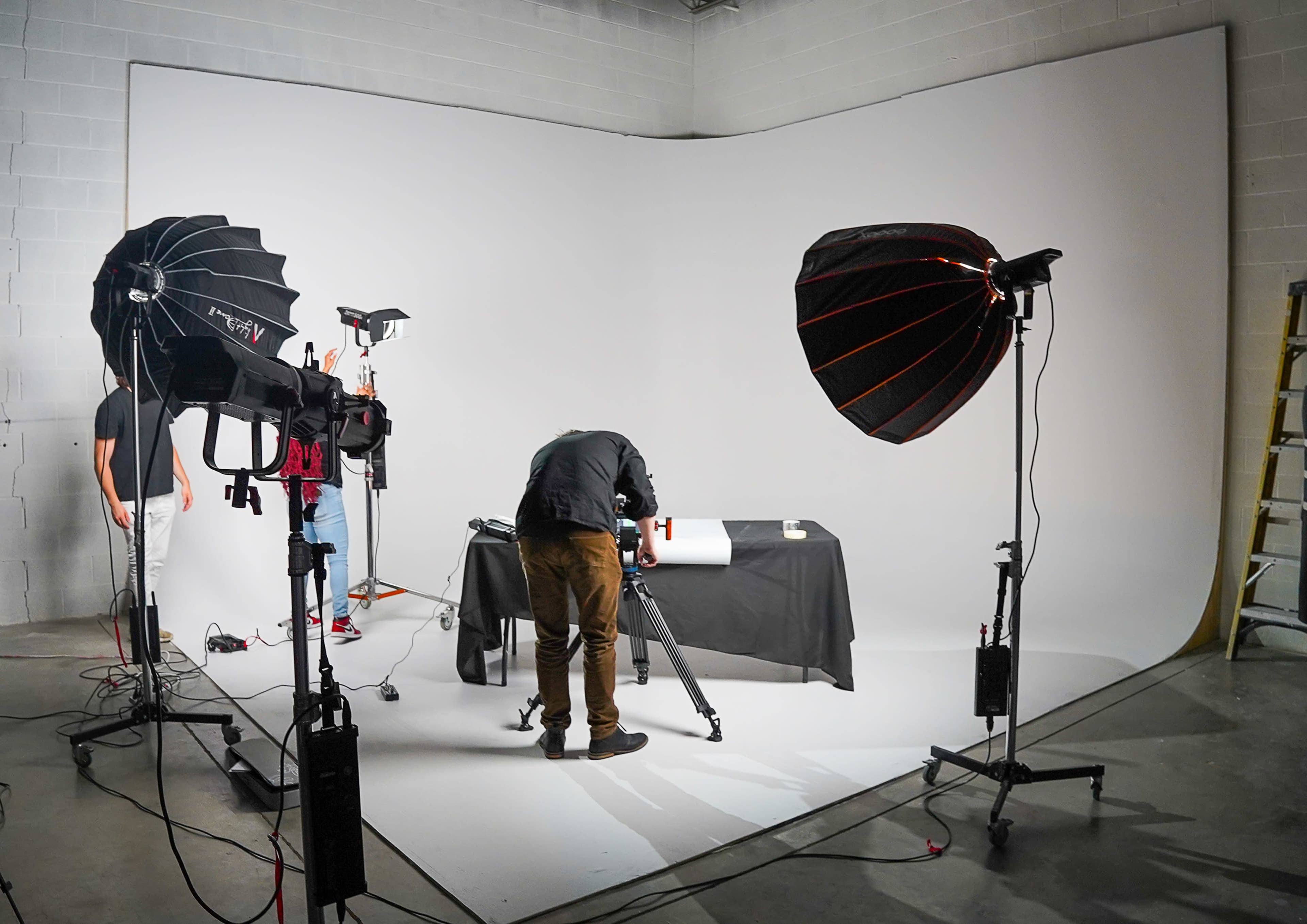 A photography studio setup features a table covered with a black cloth, surrounded by lighting equipment and a person adjusting a camera.