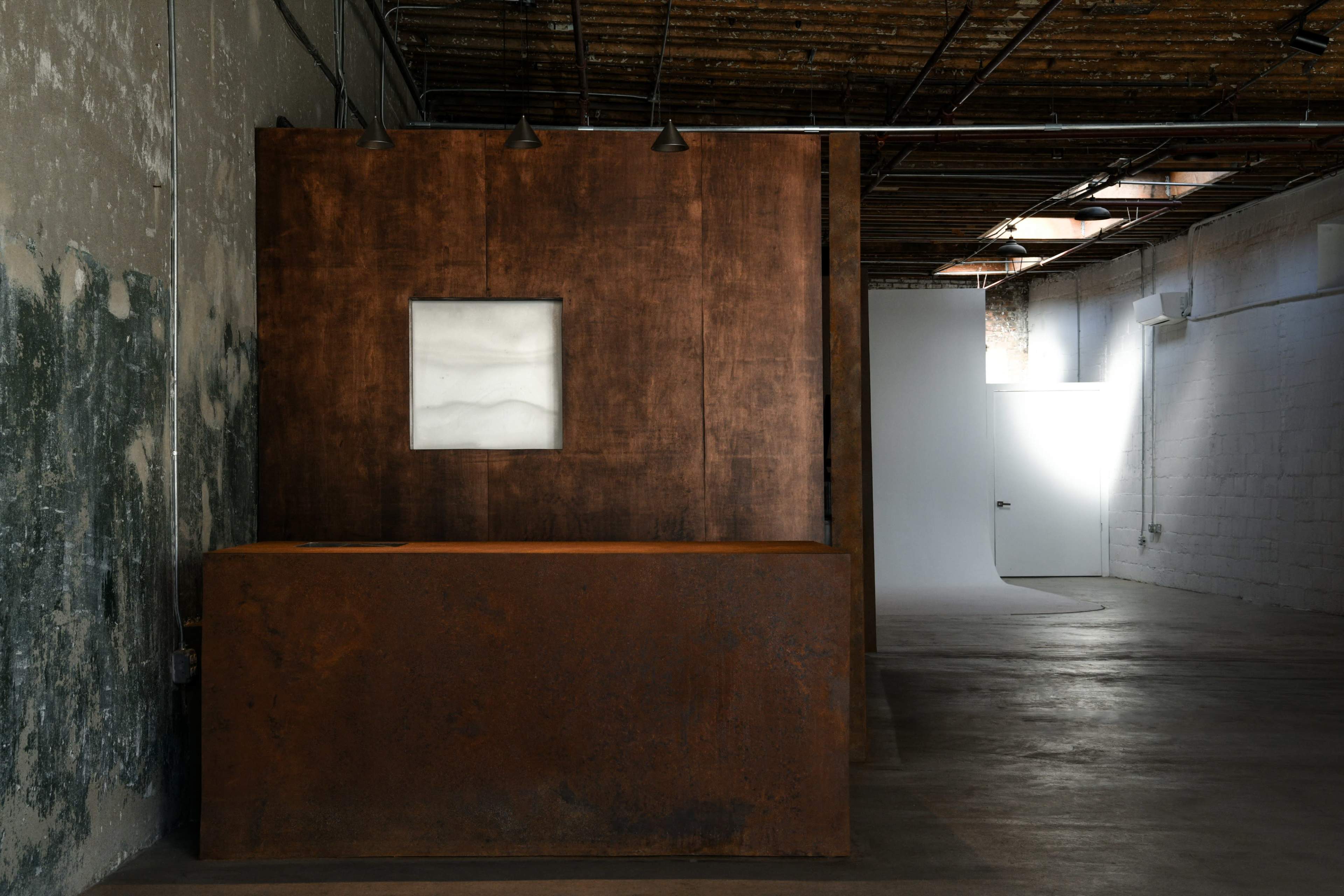 The image shows an industrial interior with a large rust-colored metal panel, featuring a square white inset, juxtaposed against a textured wall, and illuminated by overhead lights.