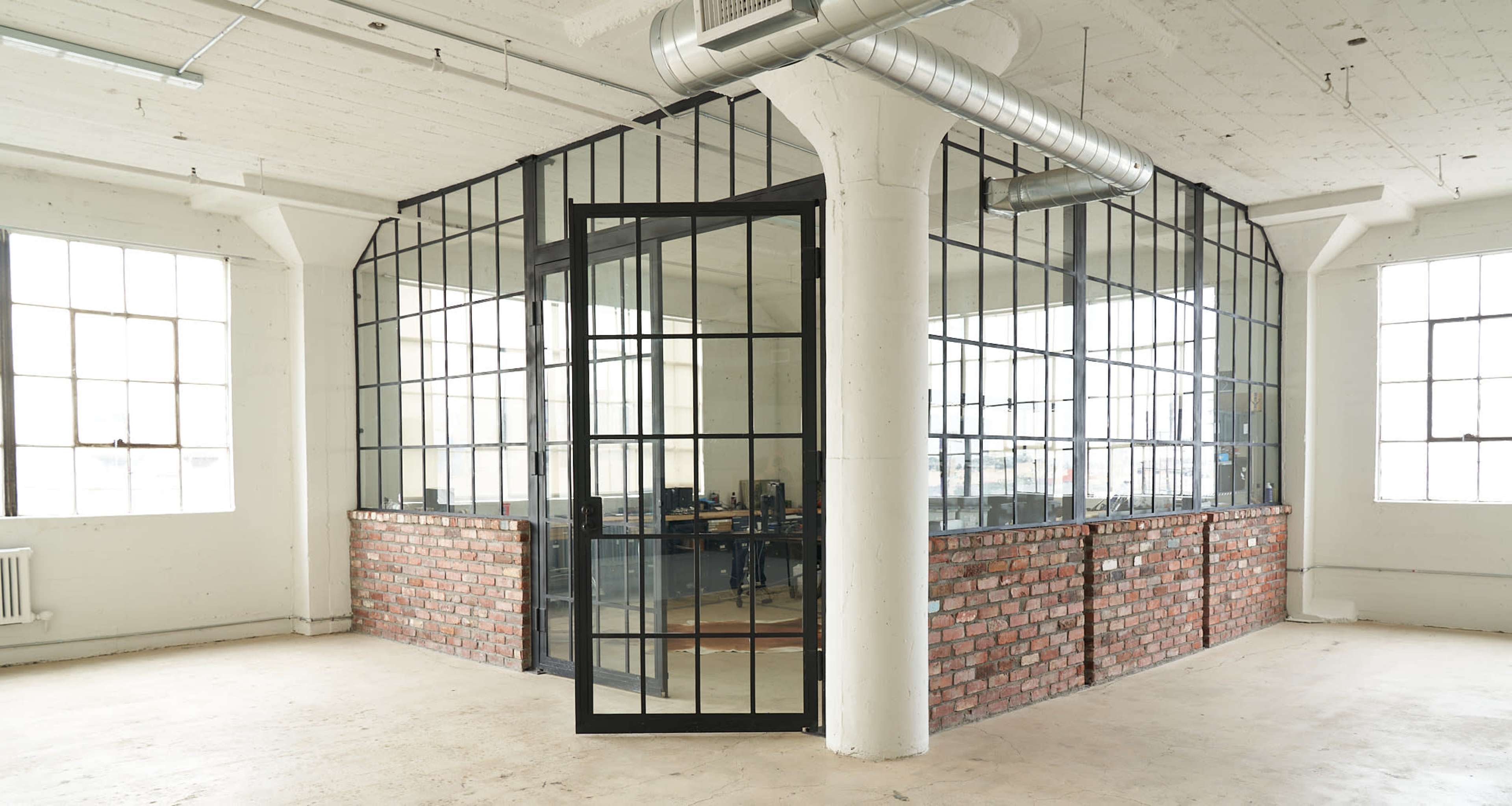 A modern office space with a glass-walled conference room featuring a brick base, large windows, and visible workstations inside.
