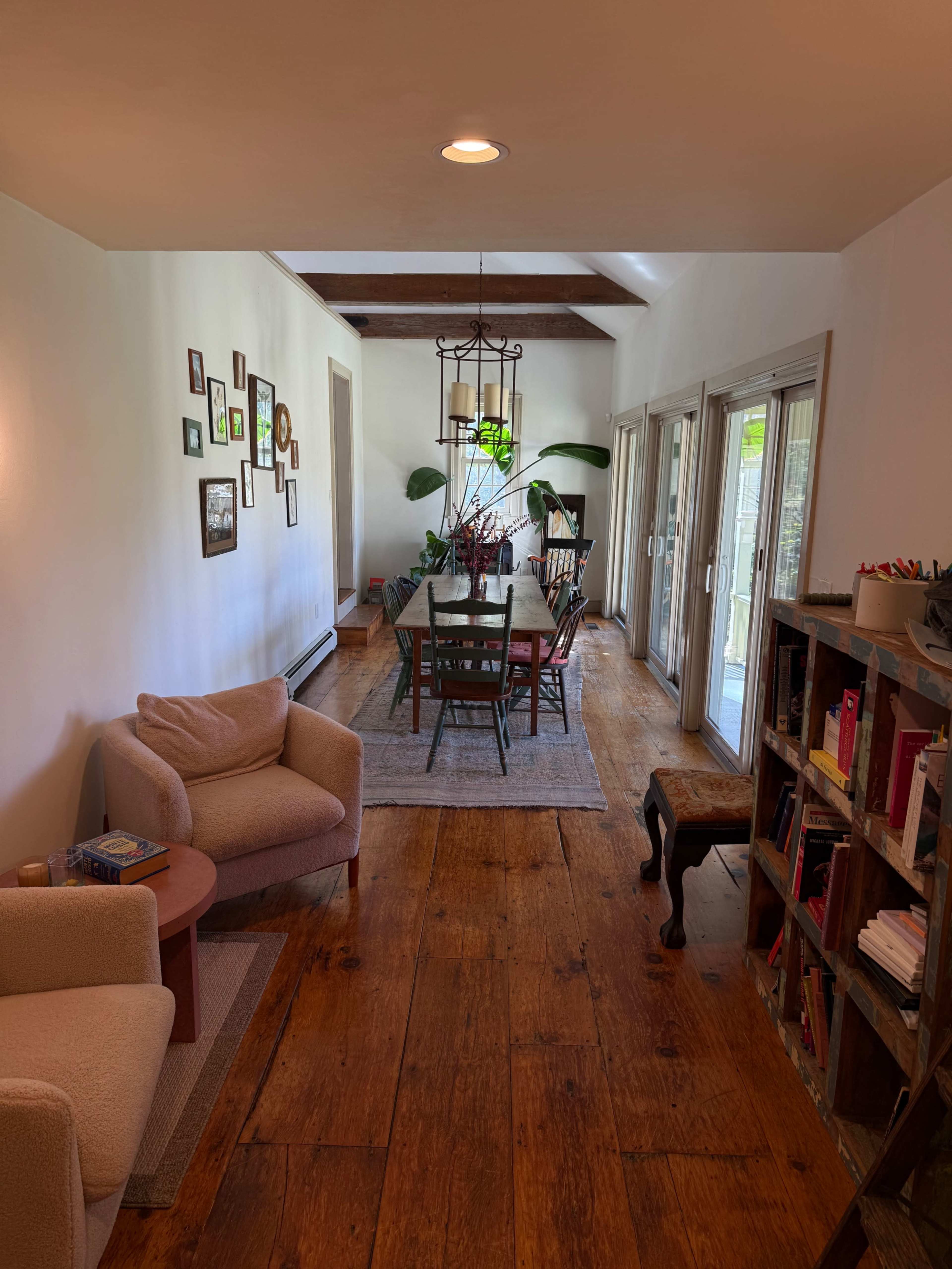 The image shows a bright, elongated dining area with a wooden table surrounded by chairs, flanked by large windows and featuring a cozy seating area in one corner.