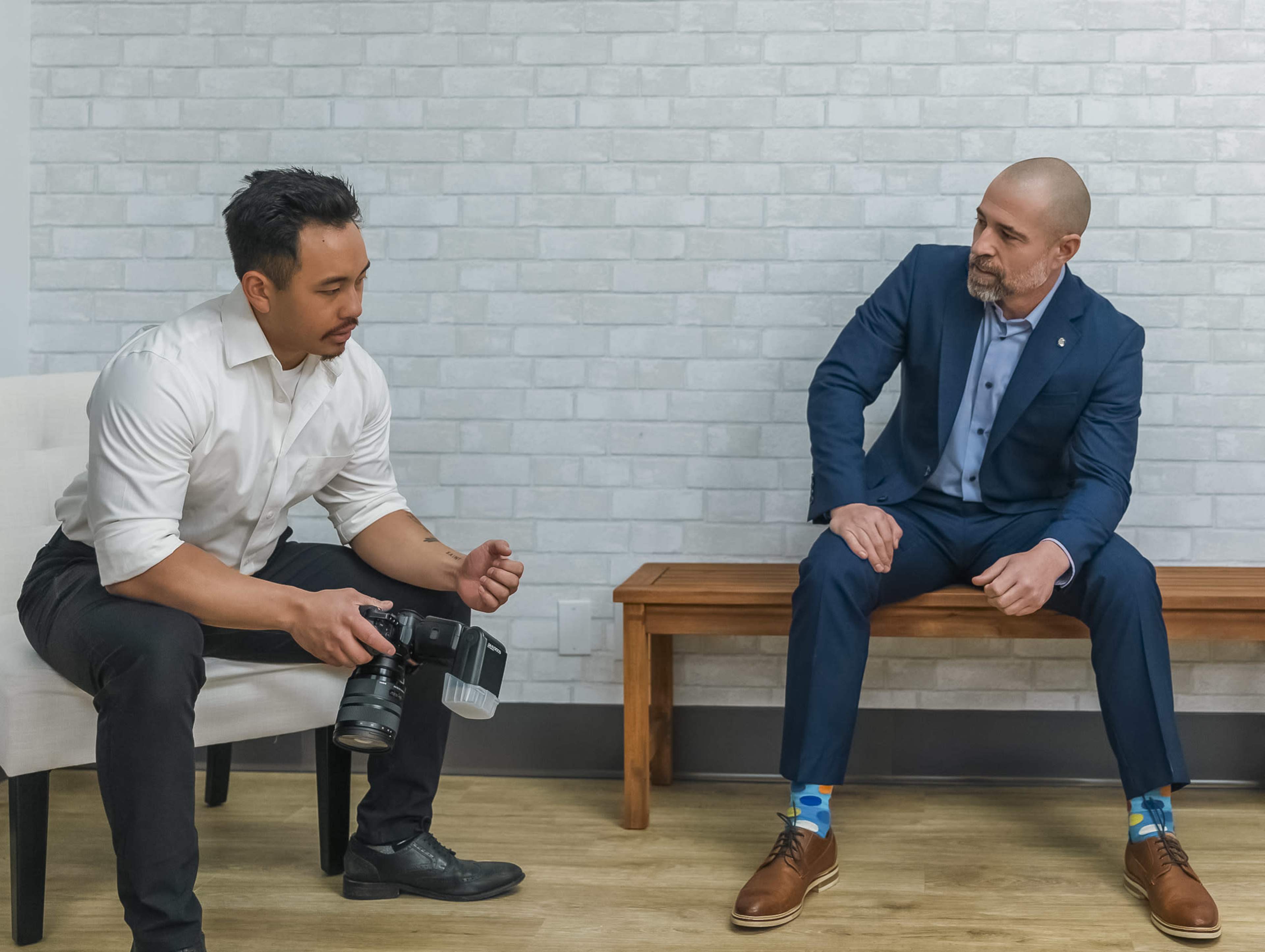 A man in a white shirt holds a camera while another man in a blue suit sits on a wooden bench, showcasing colorful socks.