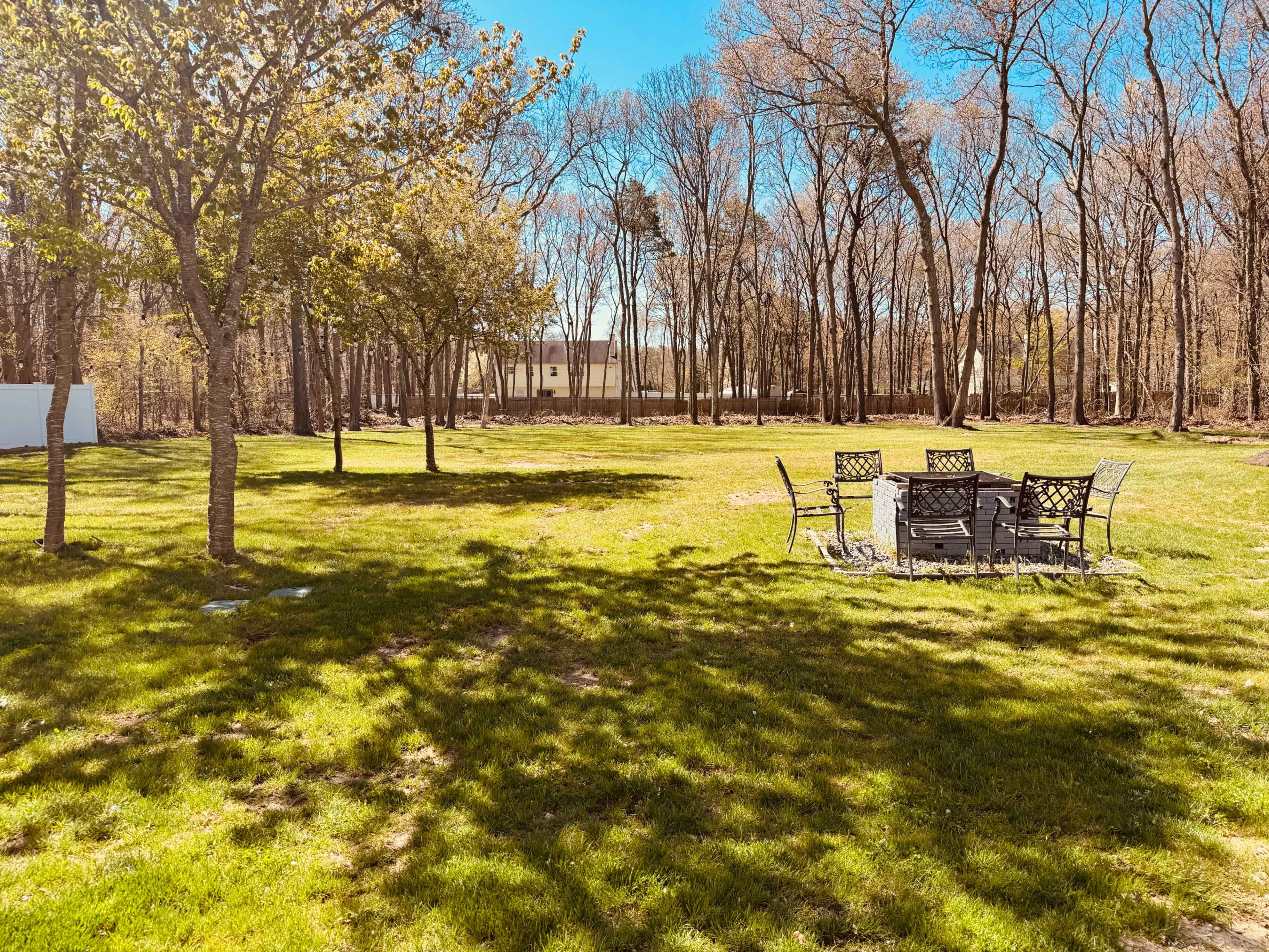 A grassy area features a metal patio set under the shade of trees, with a wooded area in the background.
