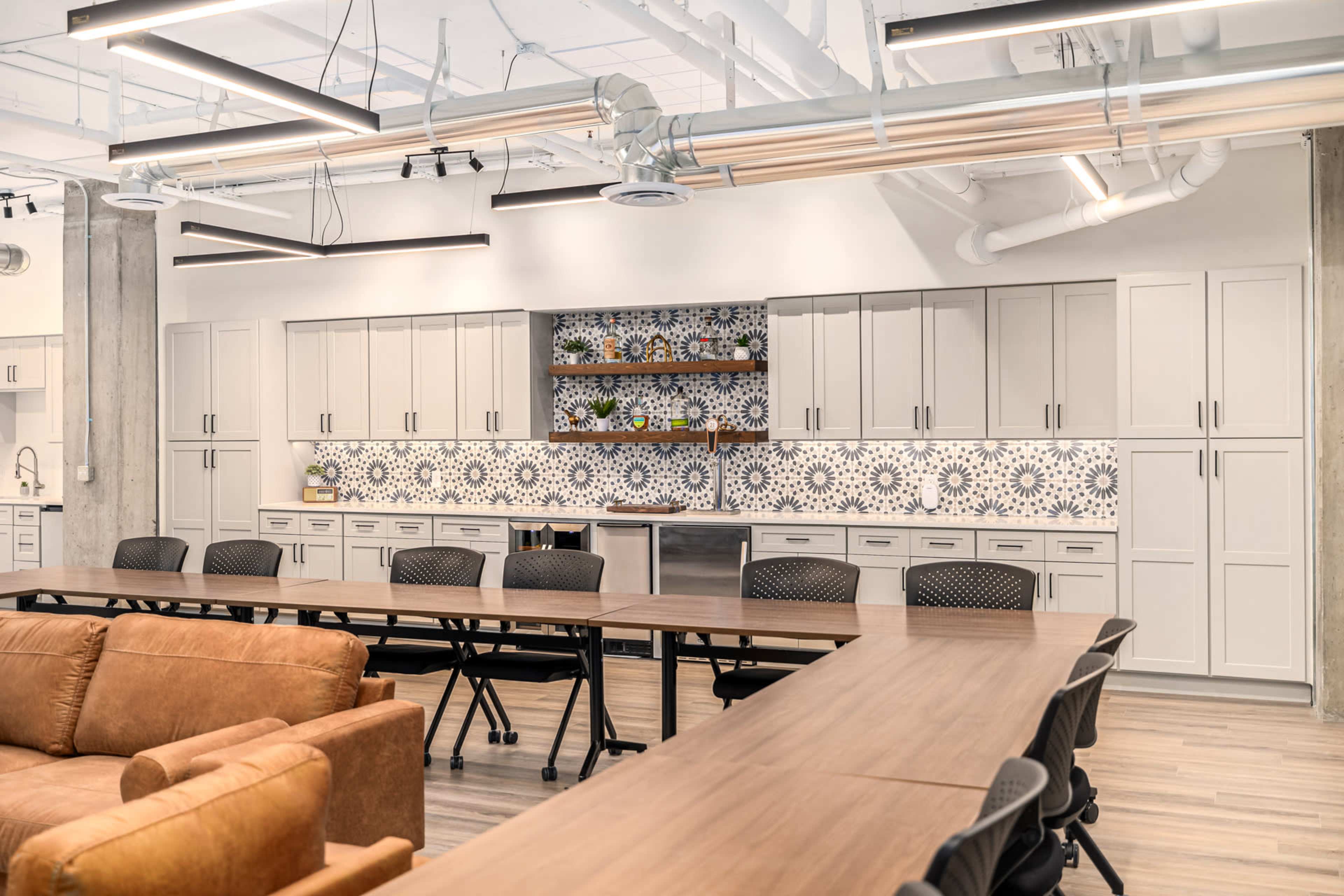 The image shows a modern communal kitchen space with a long wooden table, brown seating, and white cabinetry featuring a decorative tile backsplash.
