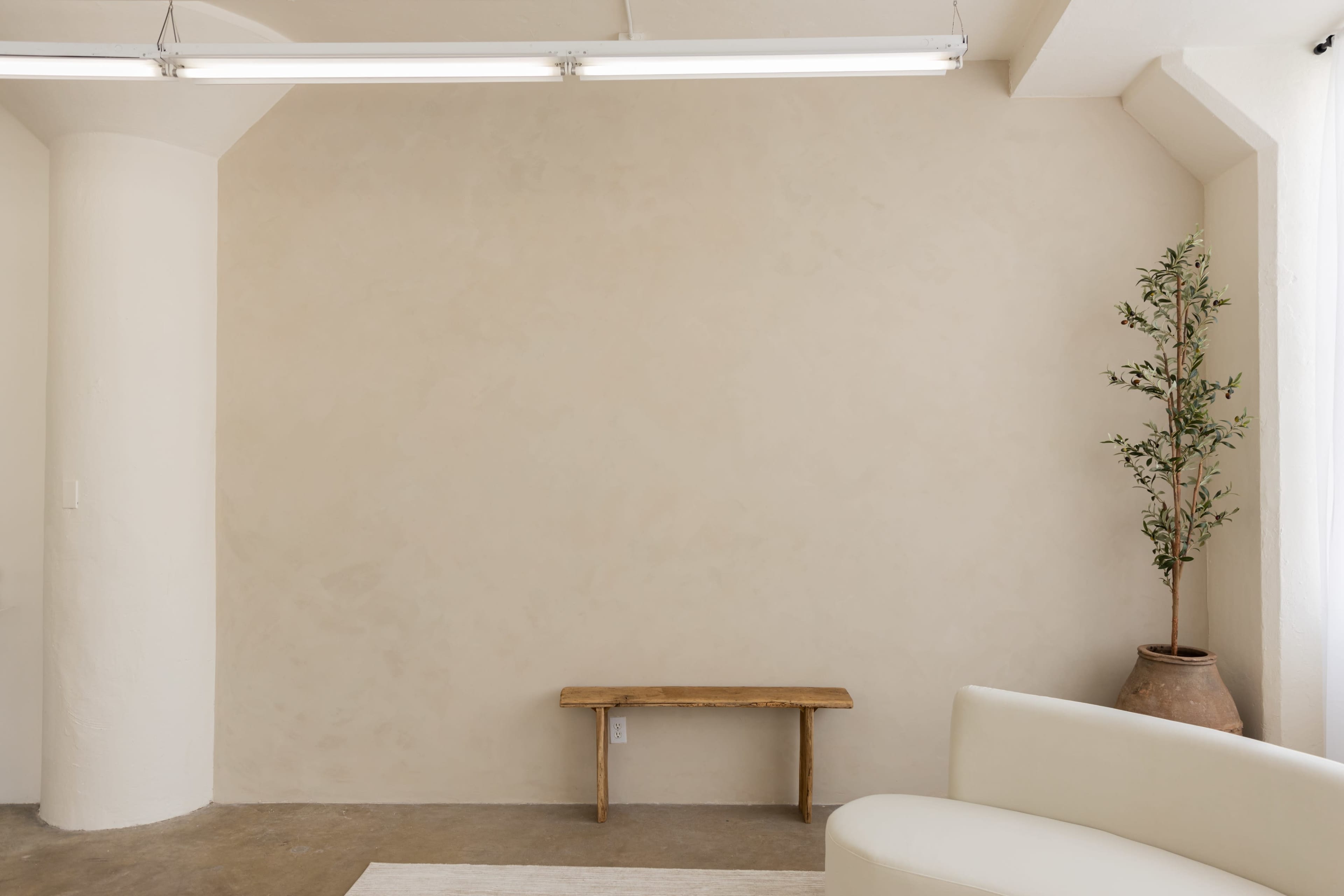 The image shows a minimalist interior with a light beige wall, a small wooden bench, a potted plant, and a white sofa.