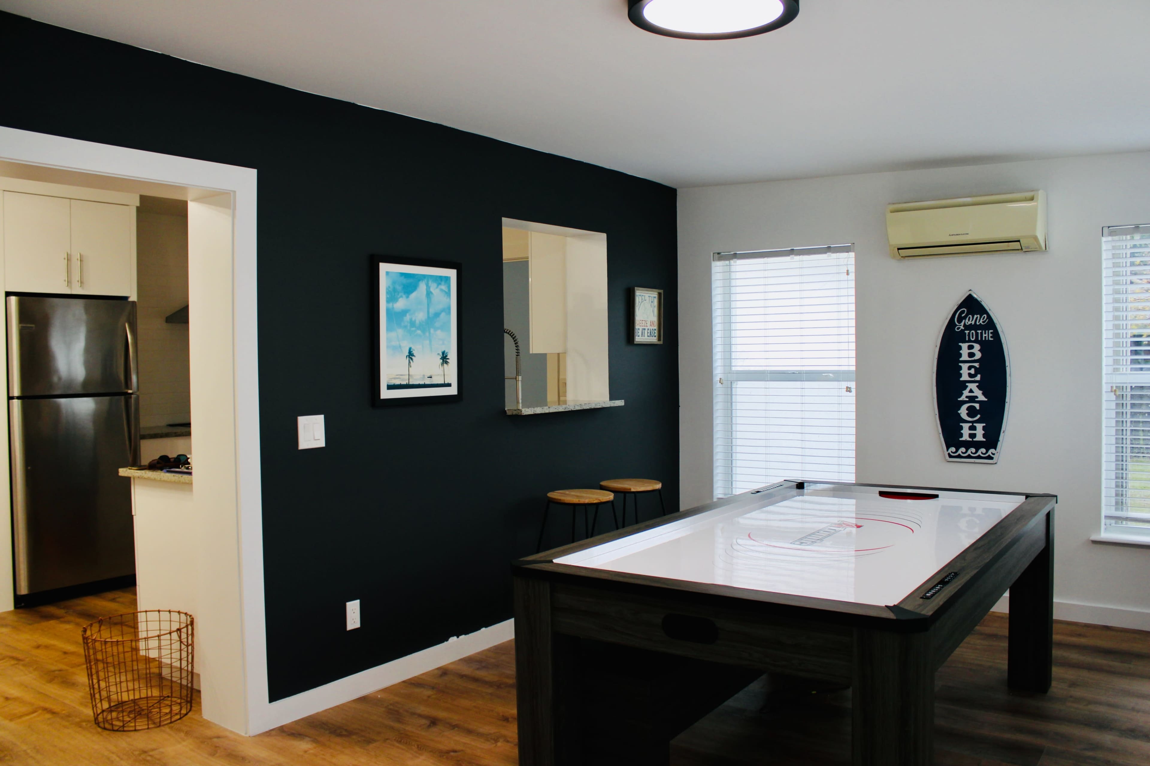 A game room features an air hockey table at the center, with a dark accent wall and an adjoining kitchenette visible through a wide opening.