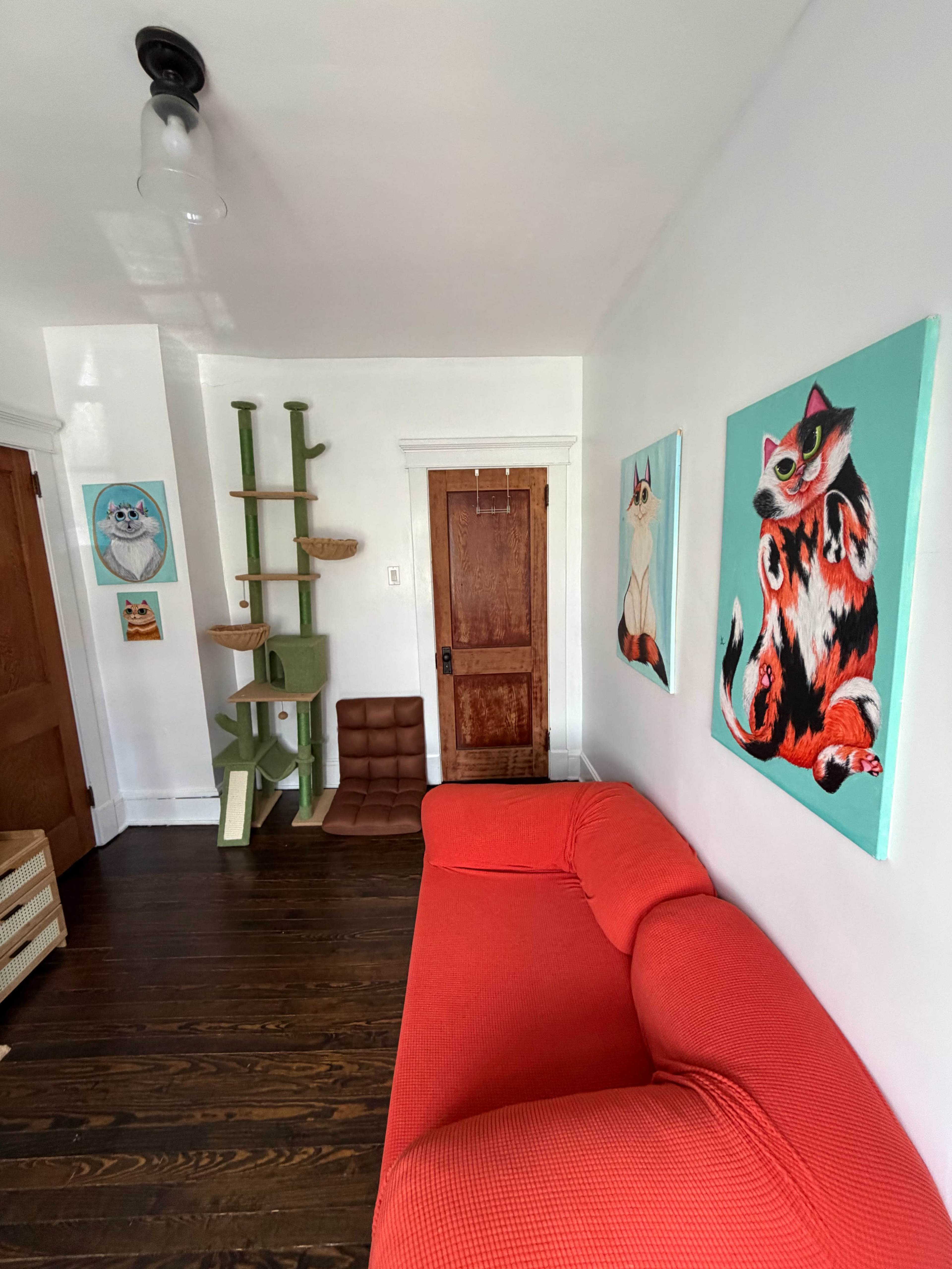 The image shows a room with a red couch, a cat tree, and paintings of cats on the walls.