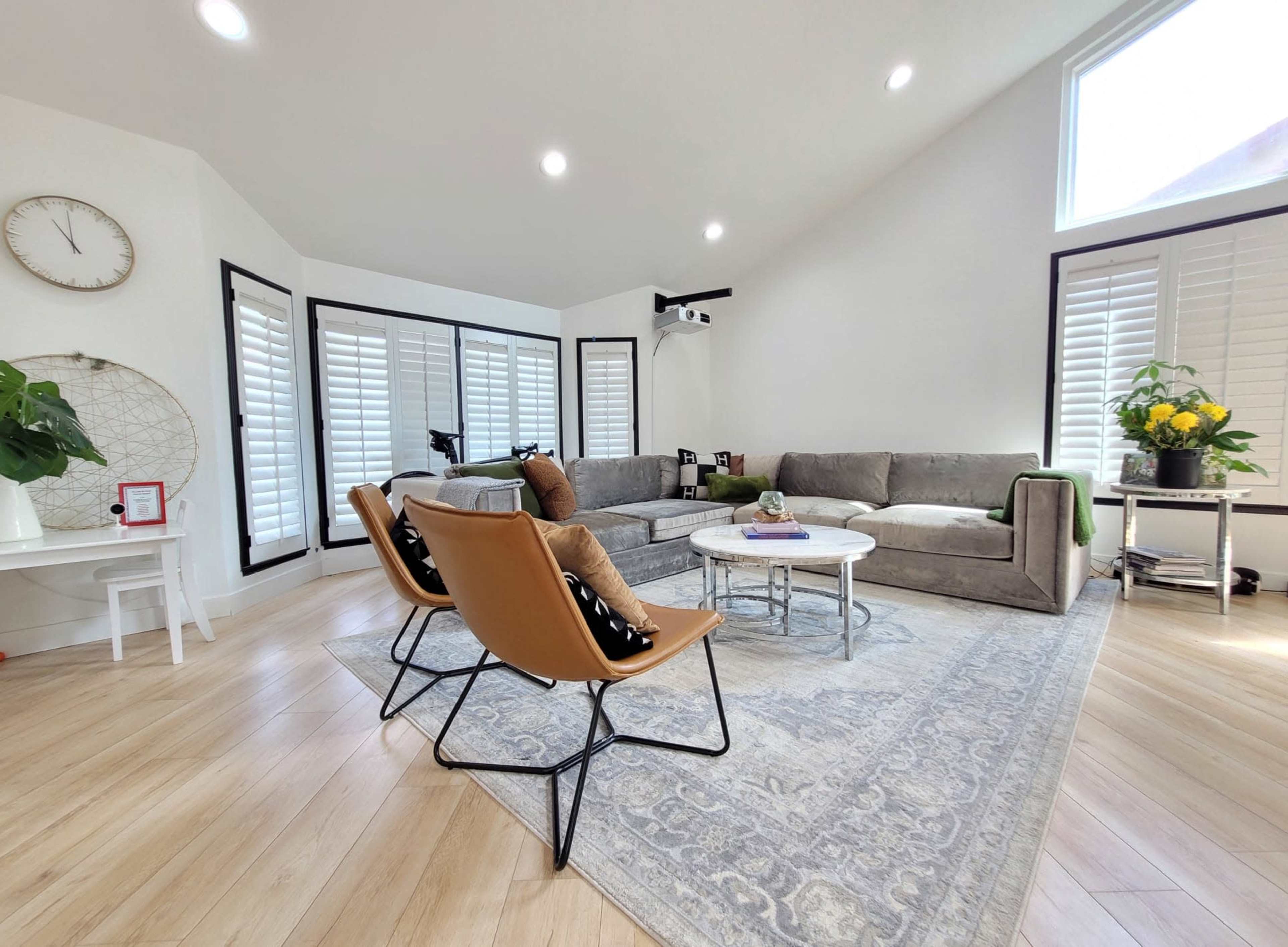 A modern living room with a gray sectional sofa, three leather chairs, a round coffee table, and large windows with plantation shutters.