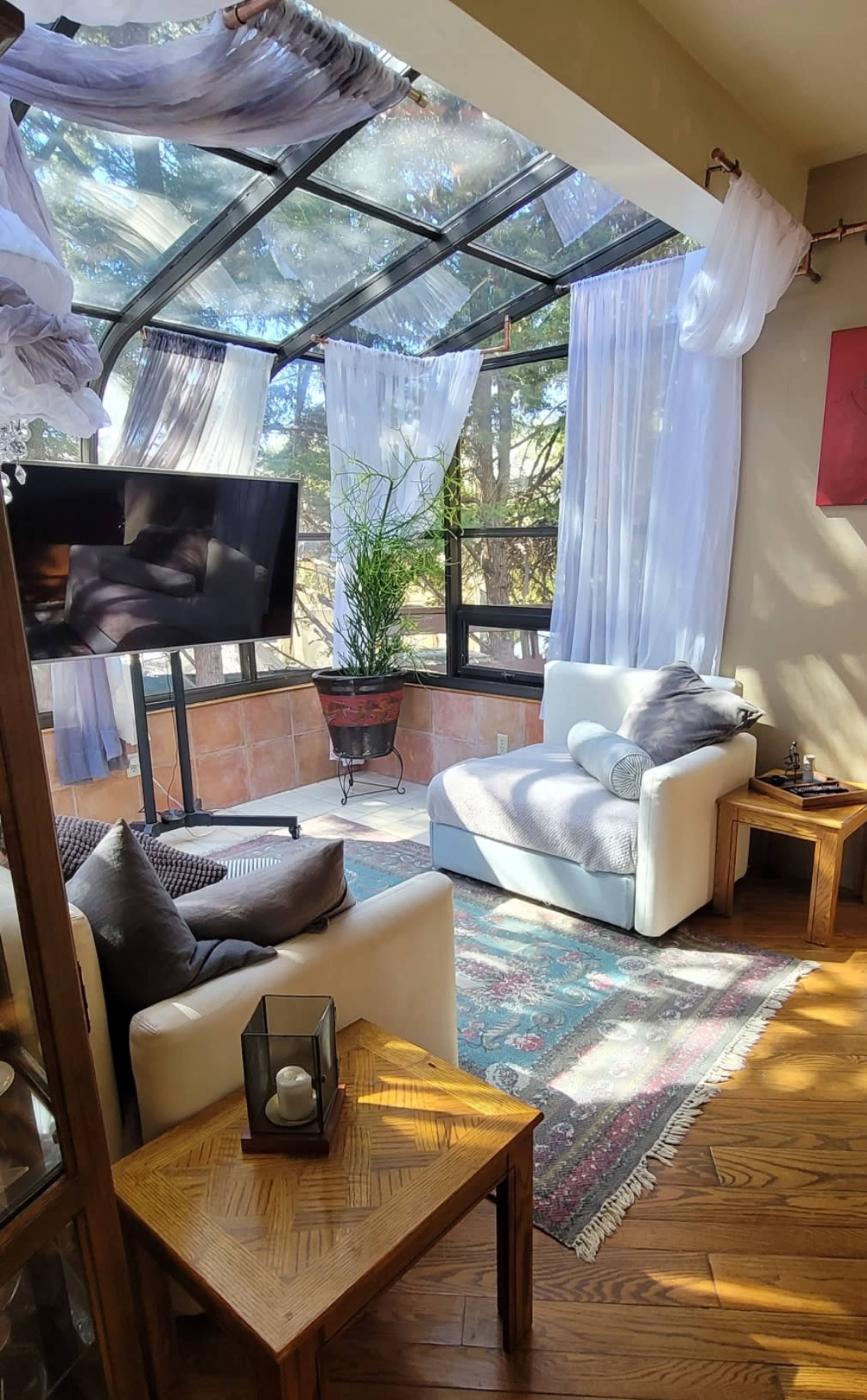 A sunlit sitting area features a glass structure with flowing drapes, a television, a comfortable couch, and a potted plant.