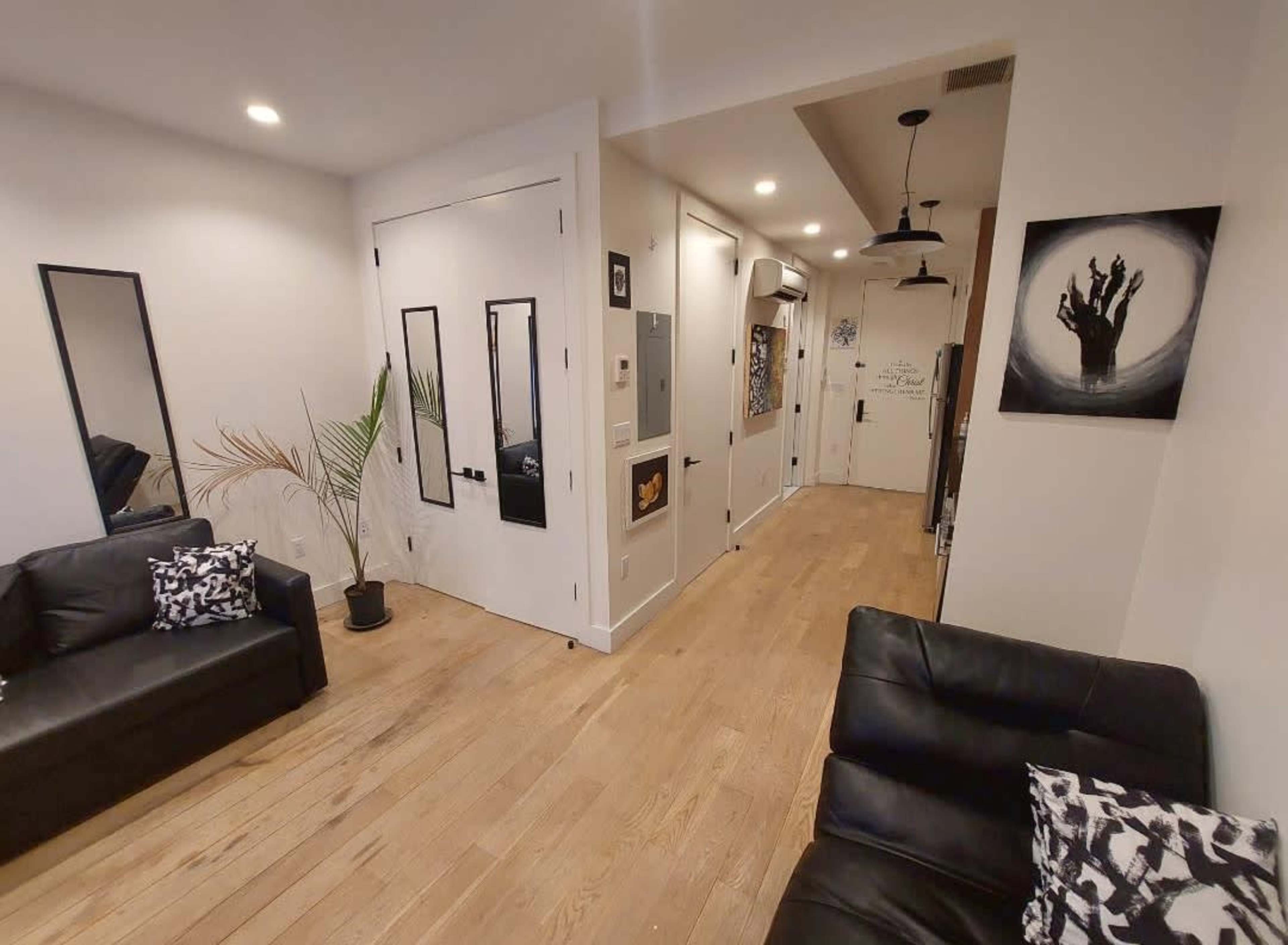 The image shows a modern living room with two black sofas, a wall-mounted artwork, and a hallway leading to a door.
