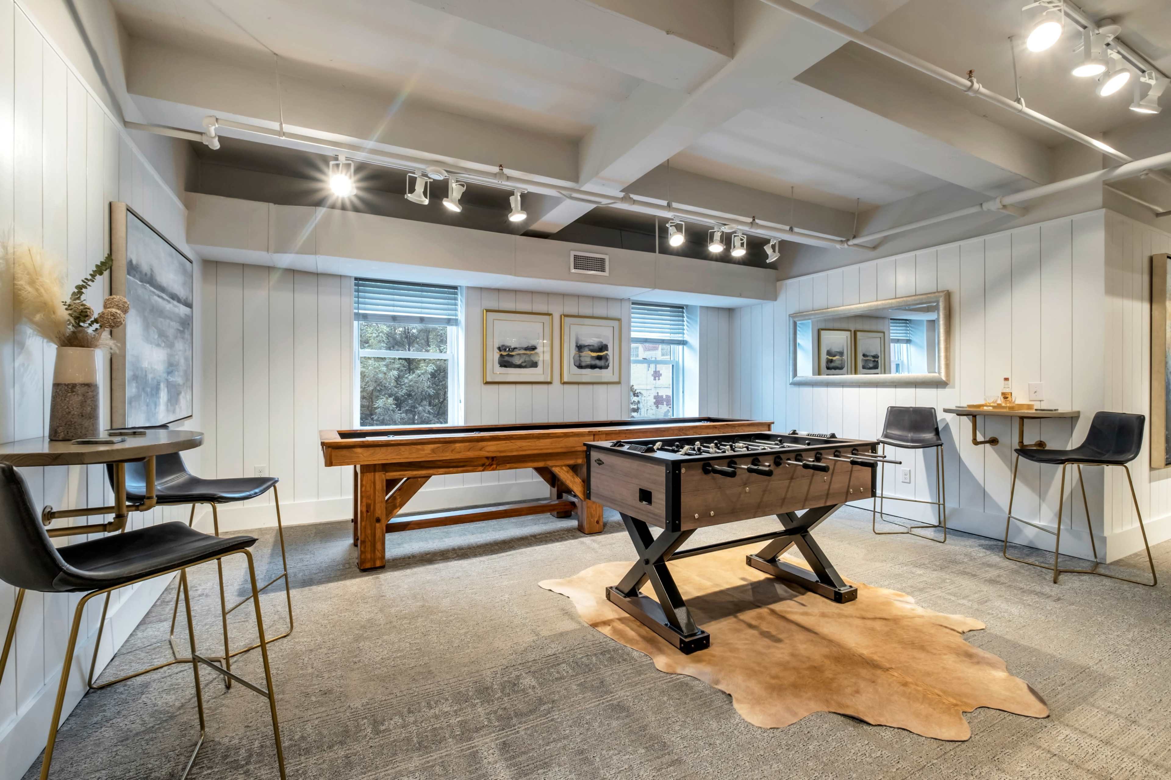 The room features a foosball table and a wooden shuffleboard along with seating and large windows that provide natural light.