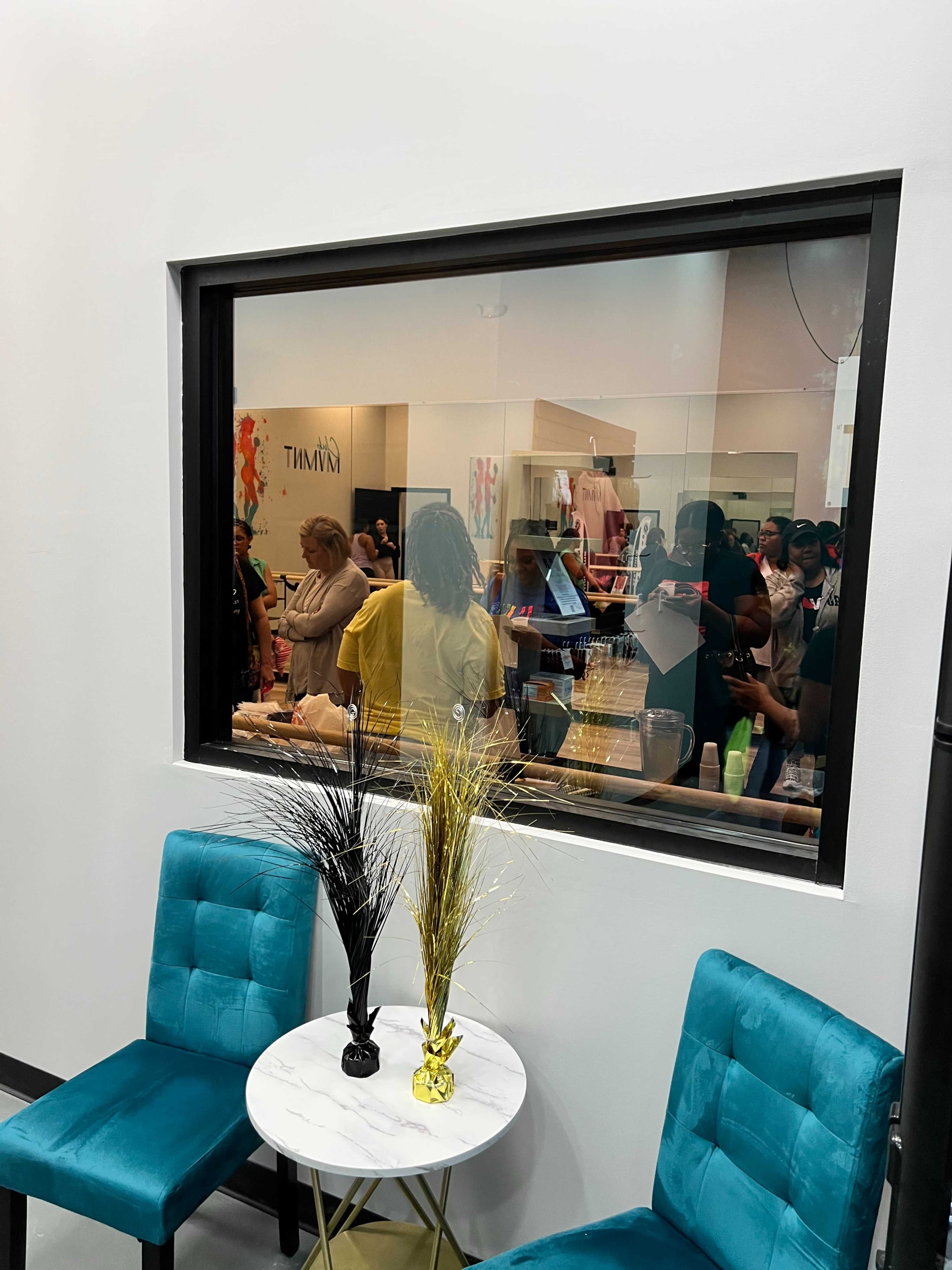 A small seating area with two teal chairs and a round table, facing a glass window through which a busy retail space can be seen filled with people and merchandise.