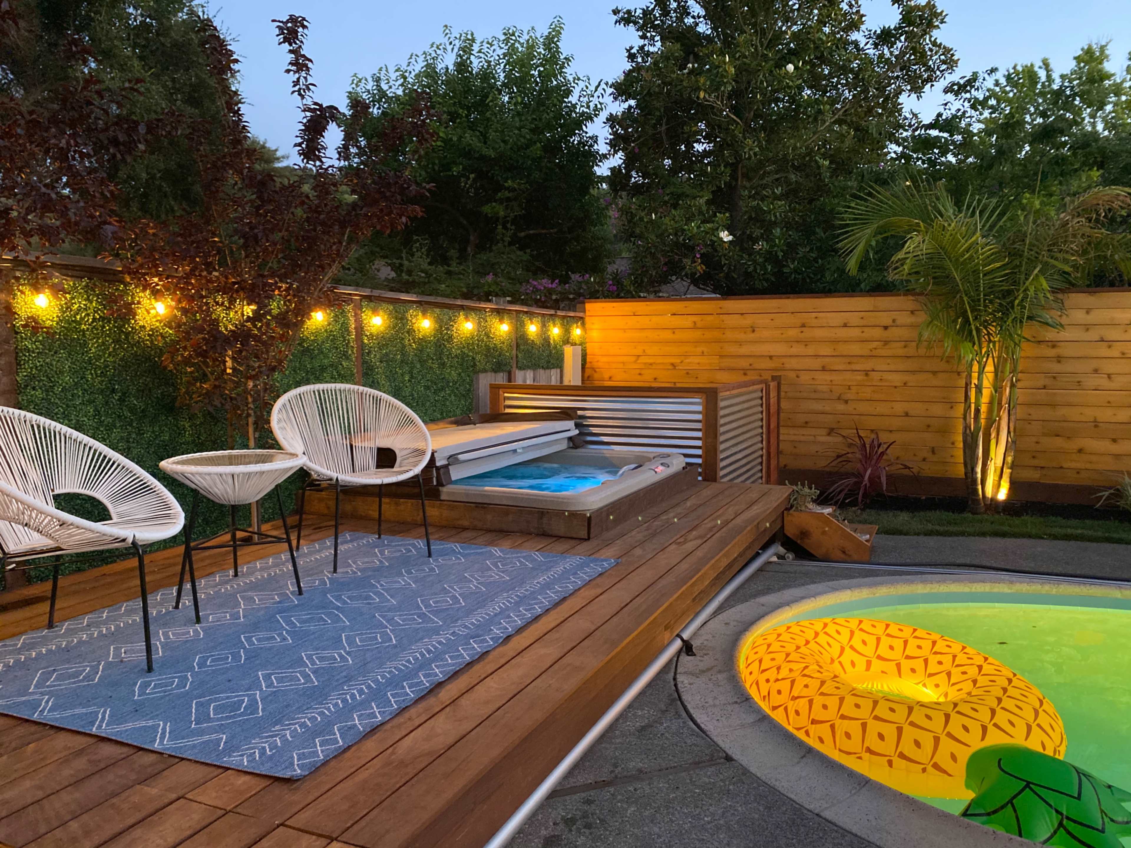 The scene shows a backyard with a wooden deck featuring two chairs, a hot tub, and a pool with a yellow pineapple design, surrounded by greenery and lit with string lights.