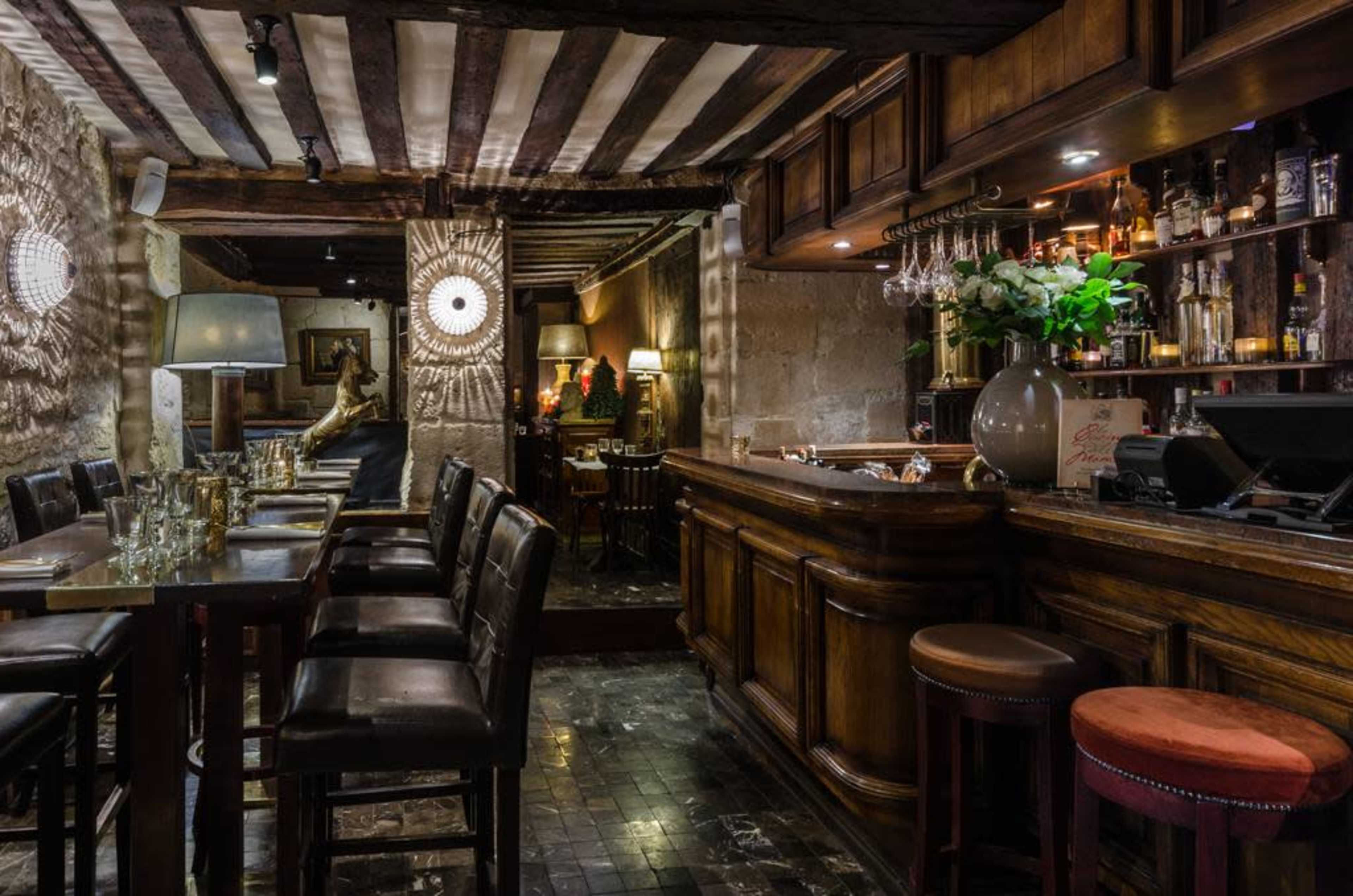 A wooden bar area with high tables and chairs, accented by decorative lighting and a variety of bottles displayed on shelves in a cozy interior.