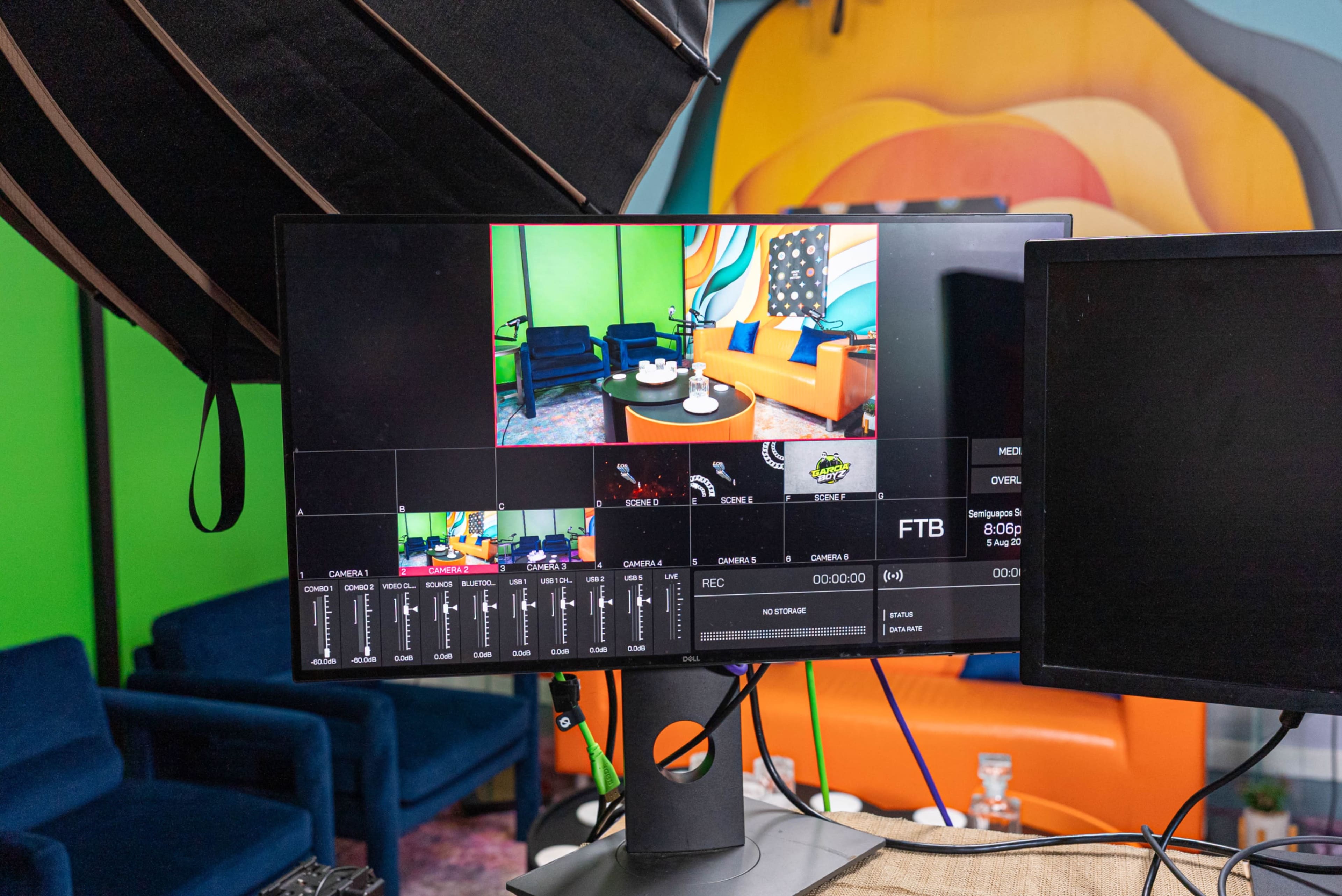 The image shows a computer monitor displaying a video production interface, with a vibrant studio background featuring colorful furniture and walls.