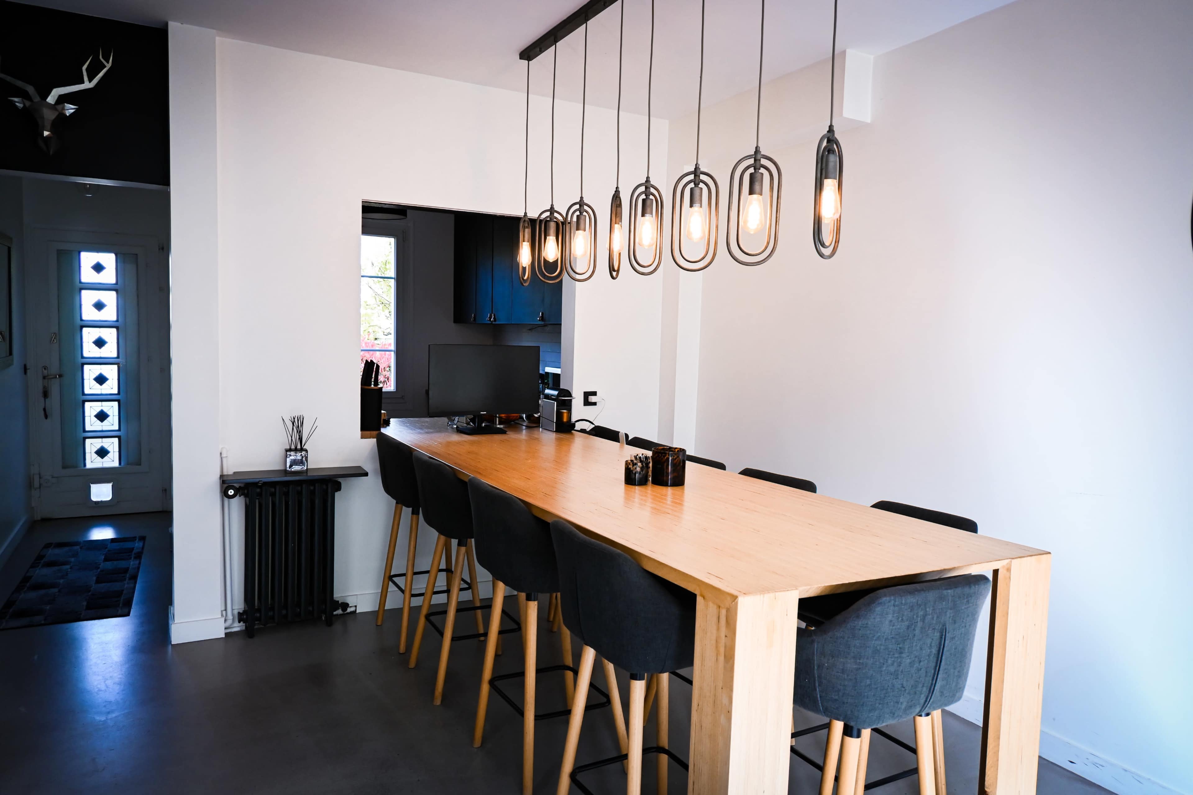 The image shows a modern dining area with a long wooden table surrounded by high stools and pendant lights hanging above it.