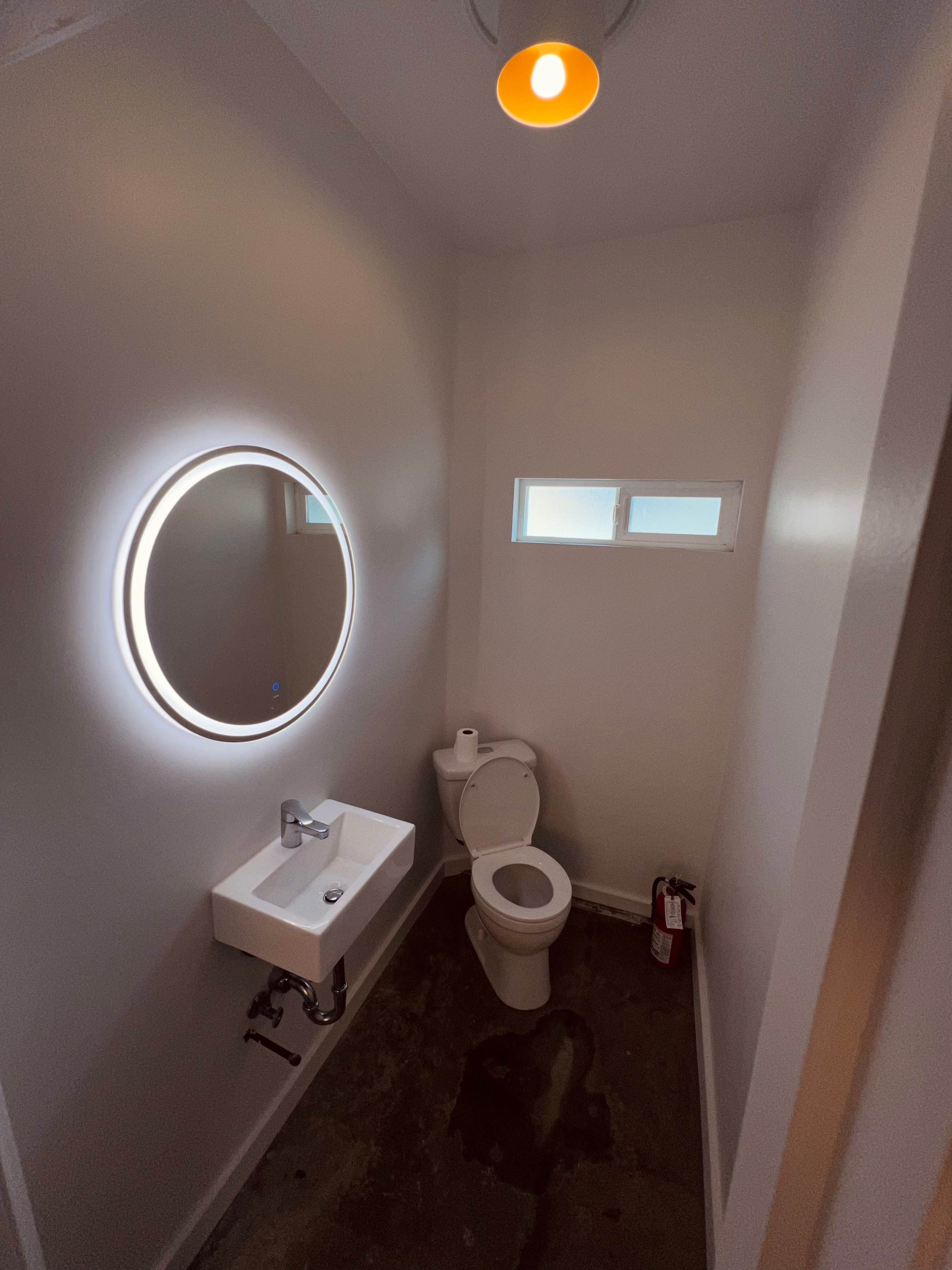 A small bathroom with a circular mirror, a white sink, a toilet, and a fire extinguisher against a plain wall.