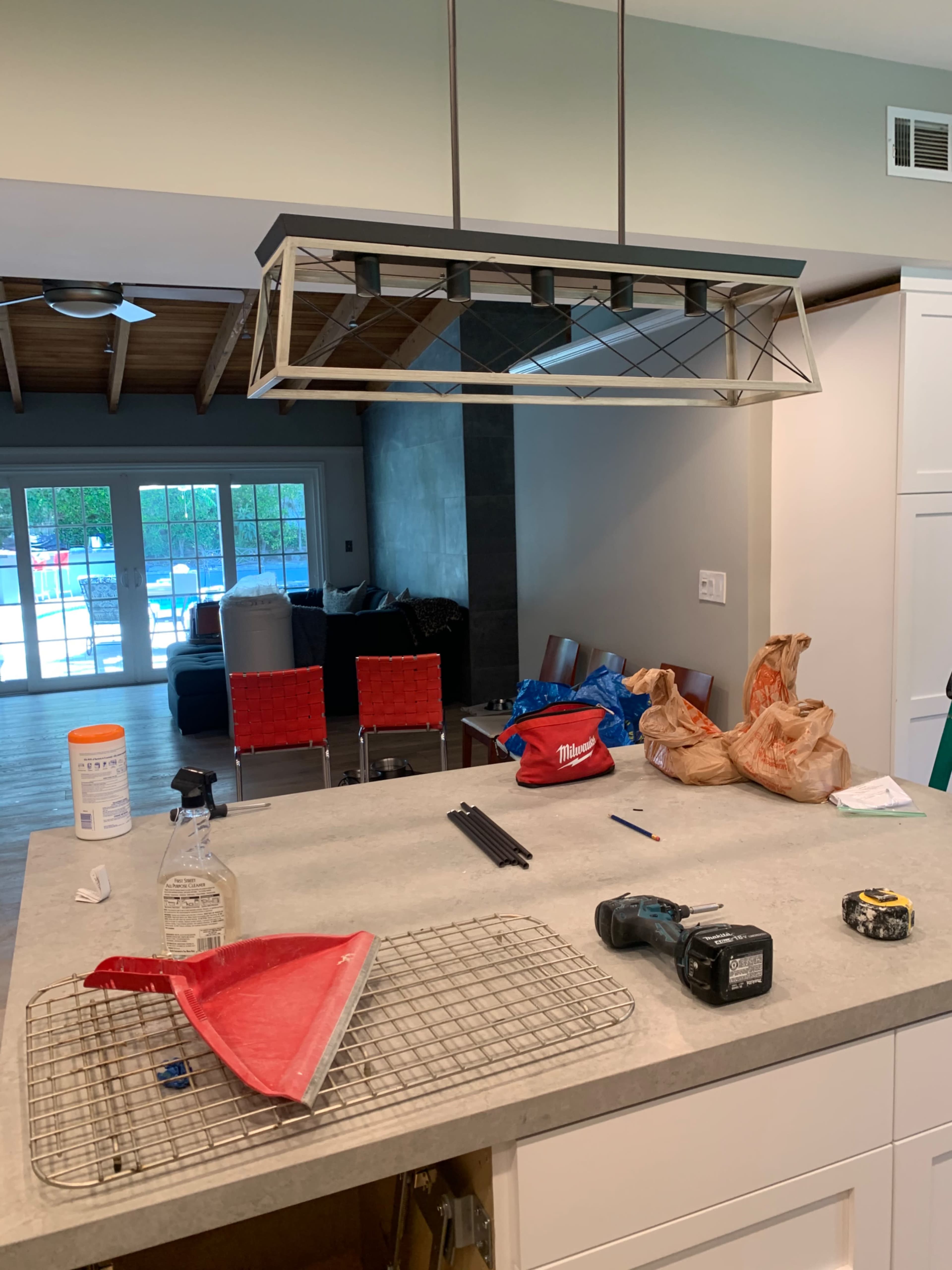 The image shows a kitchen with a pendant light fixture above a countertop cluttered with cleaning supplies, tools, and bags, while a living area with red chairs and large windows is visible in the background.