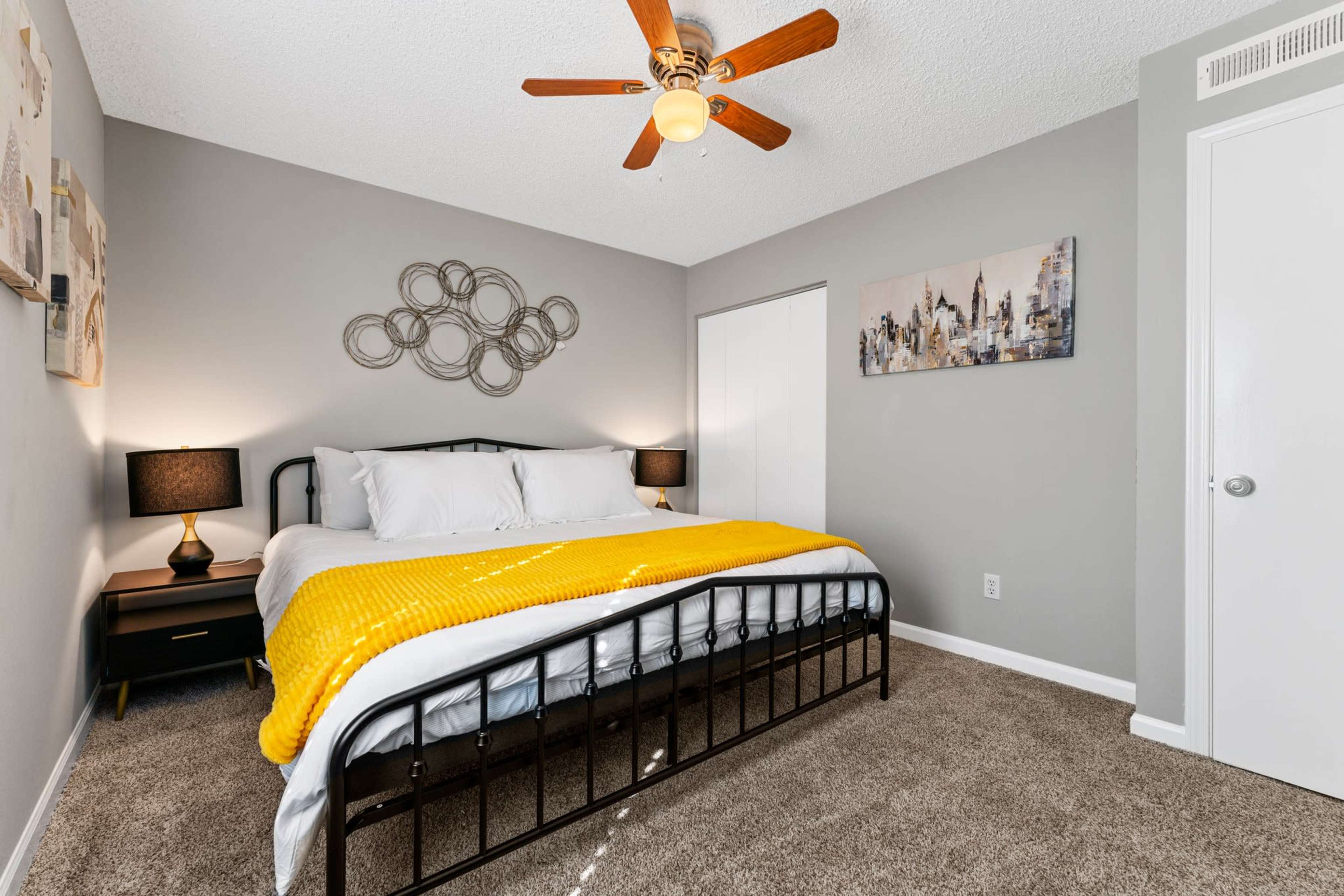 A neatly arranged bedroom featuring a black metal bed with white bedding and a yellow throw blanket, complemented by a ceiling fan and modern artwork on the walls.