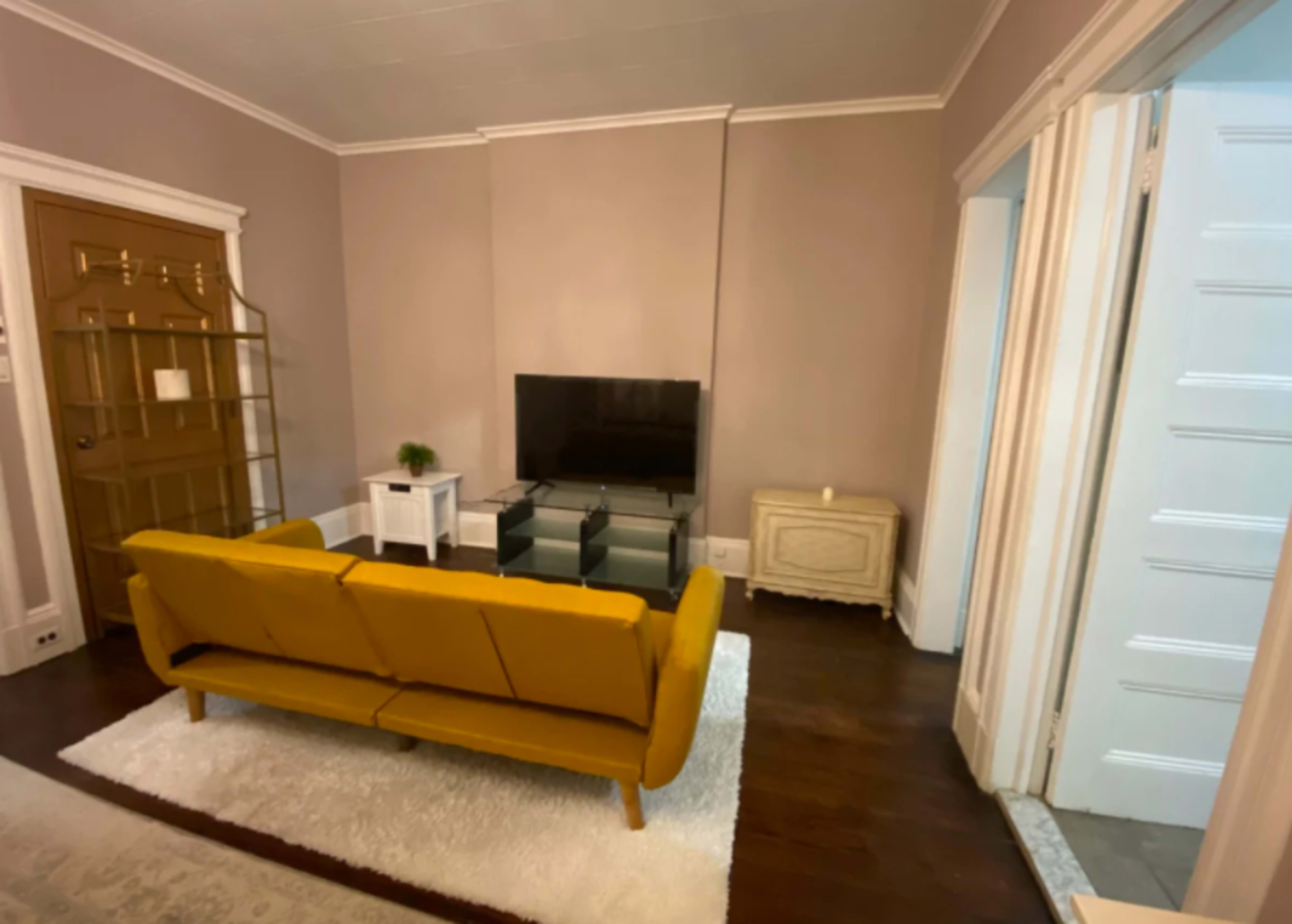 The image shows a living room with a mustard yellow sofa facing a wall-mounted television over a glass media stand, flanked by a door and a hallway.