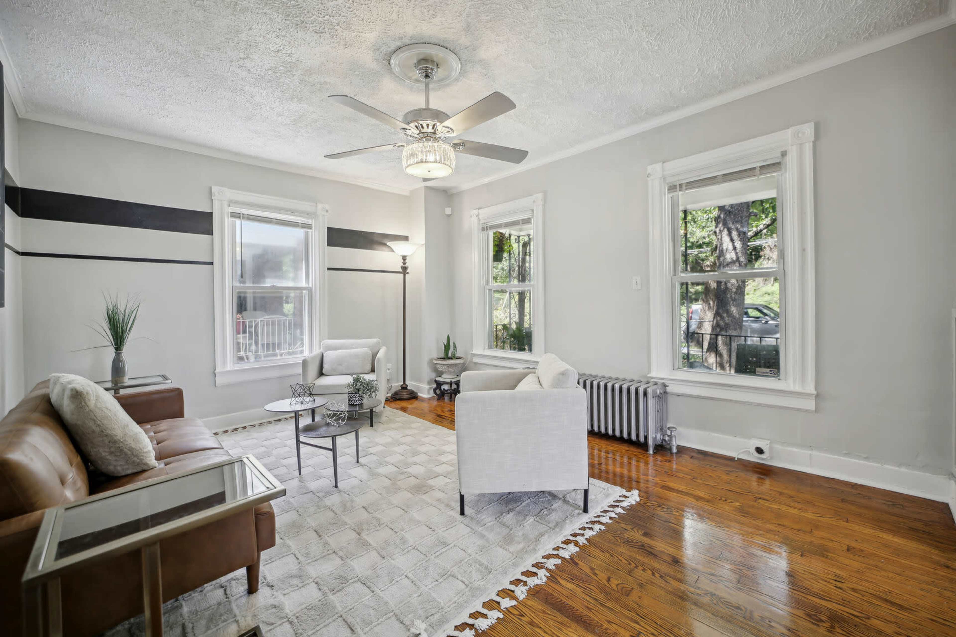 A living room features two armchairs and a leather sofa, with large windows and a ceiling fan above.