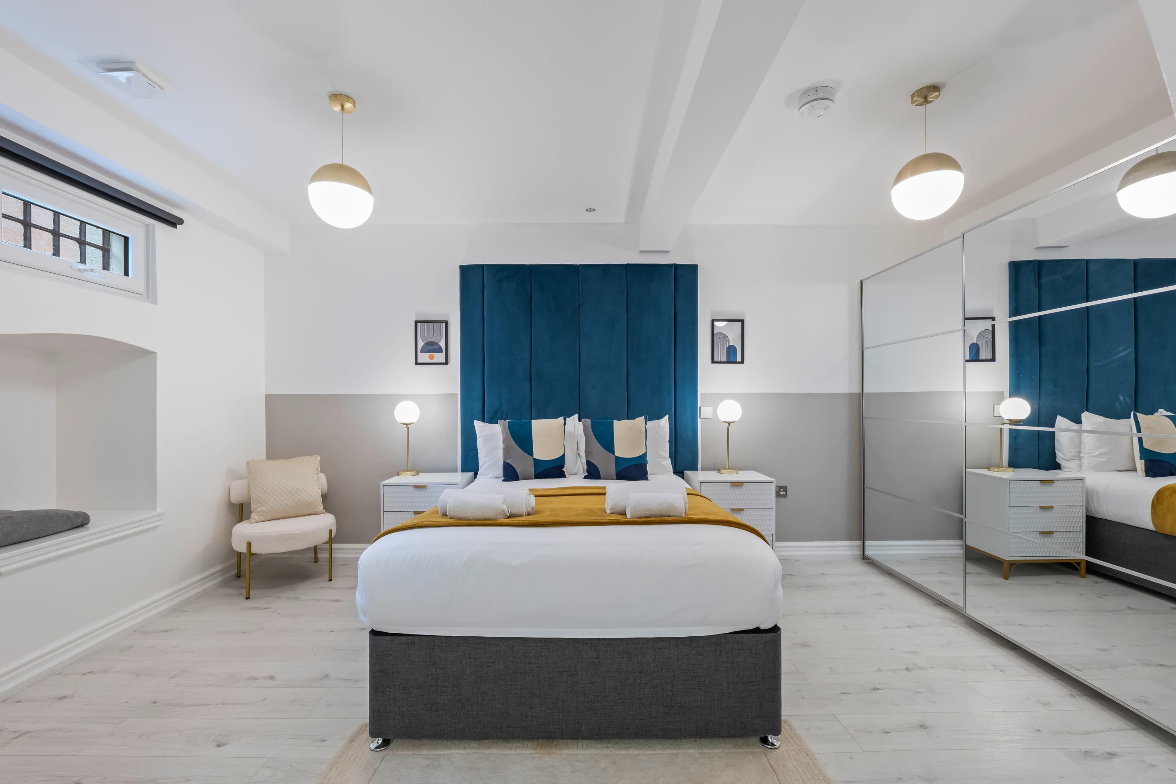 The image shows a modern bedroom featuring a bed with a gray base, blue accent wall behind the headboard, and two pendant lights above nightstands with lamps.