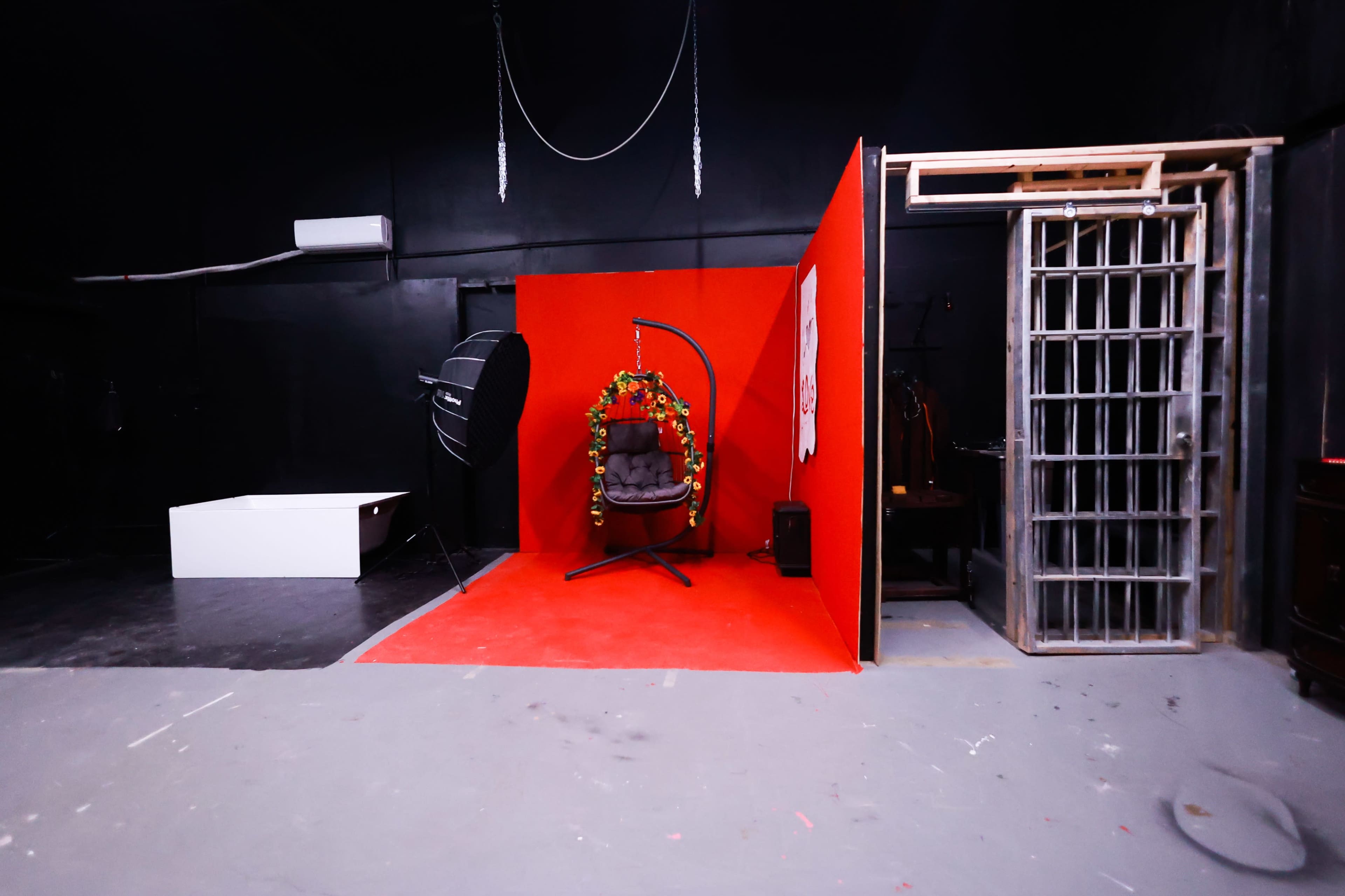 A studio space featuring a red backdrop with a hanging chair decorated with foliage, a lighting setup, and a metal cage-like structure.