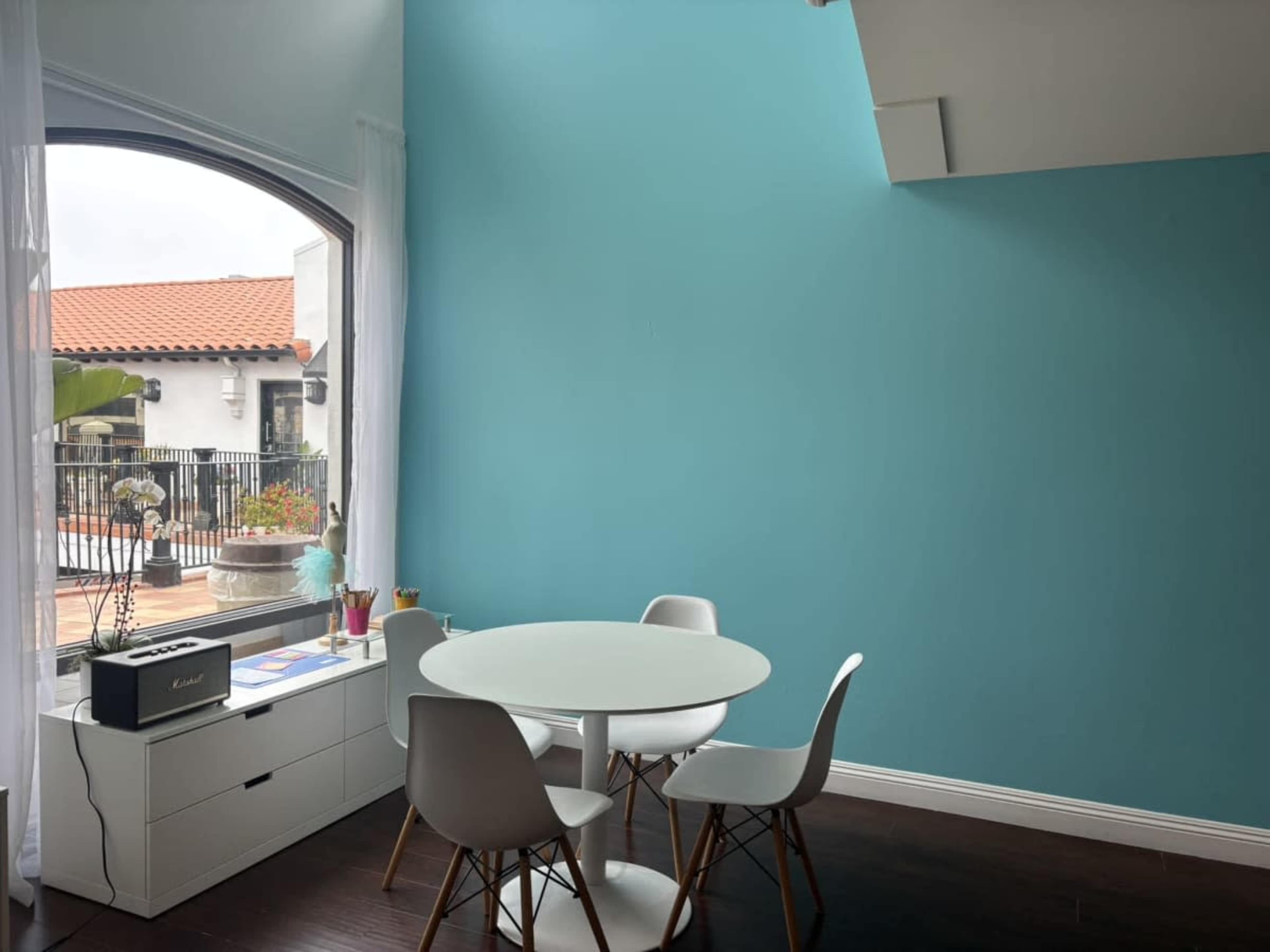 A modern dining area features a round white table surrounded by four white chairs, against a teal wall with a large window showing an outdoor patio.