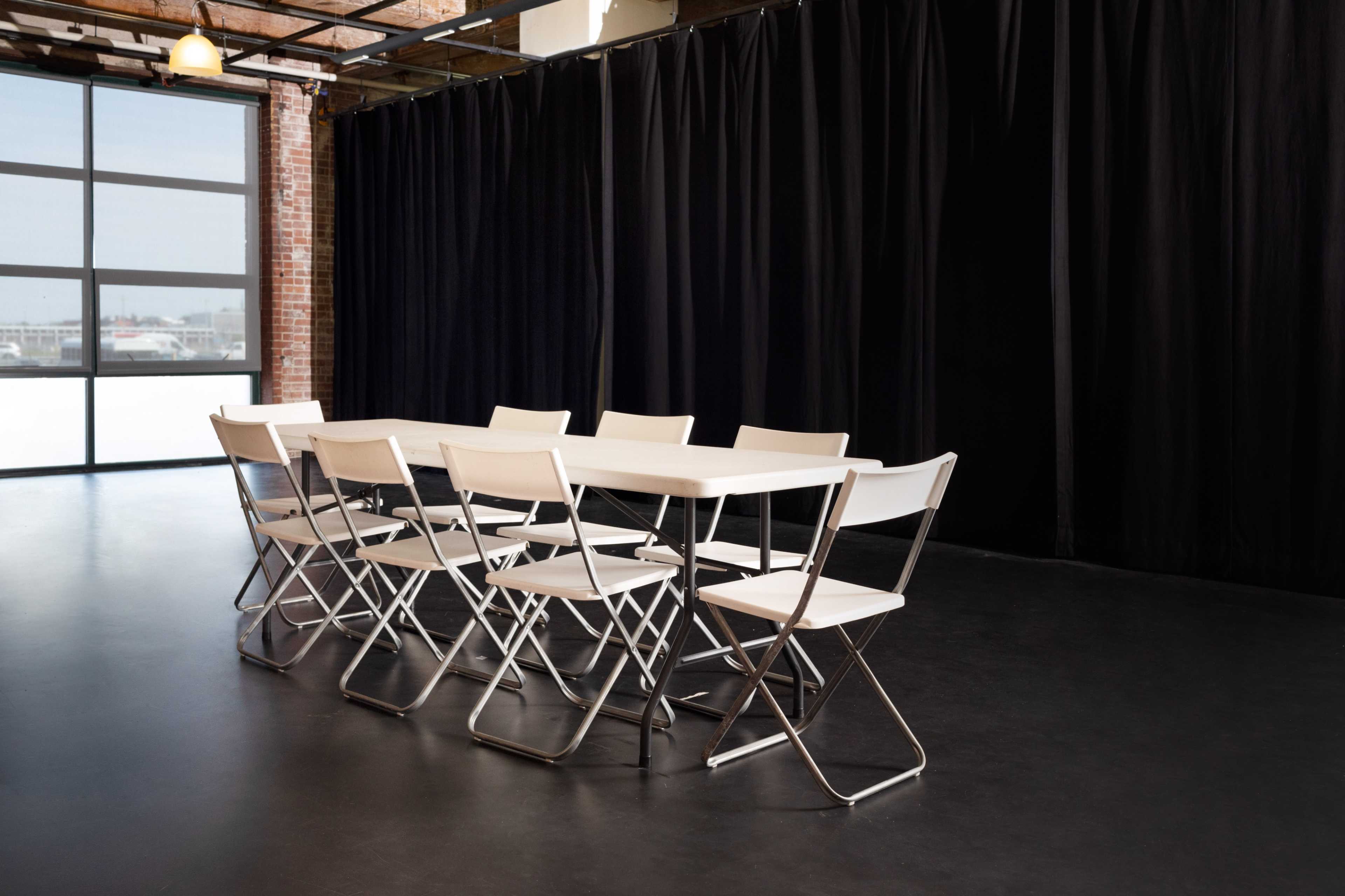 A long, white foldable table is arranged with eight matching chairs in a room featuring large windows and black curtains.