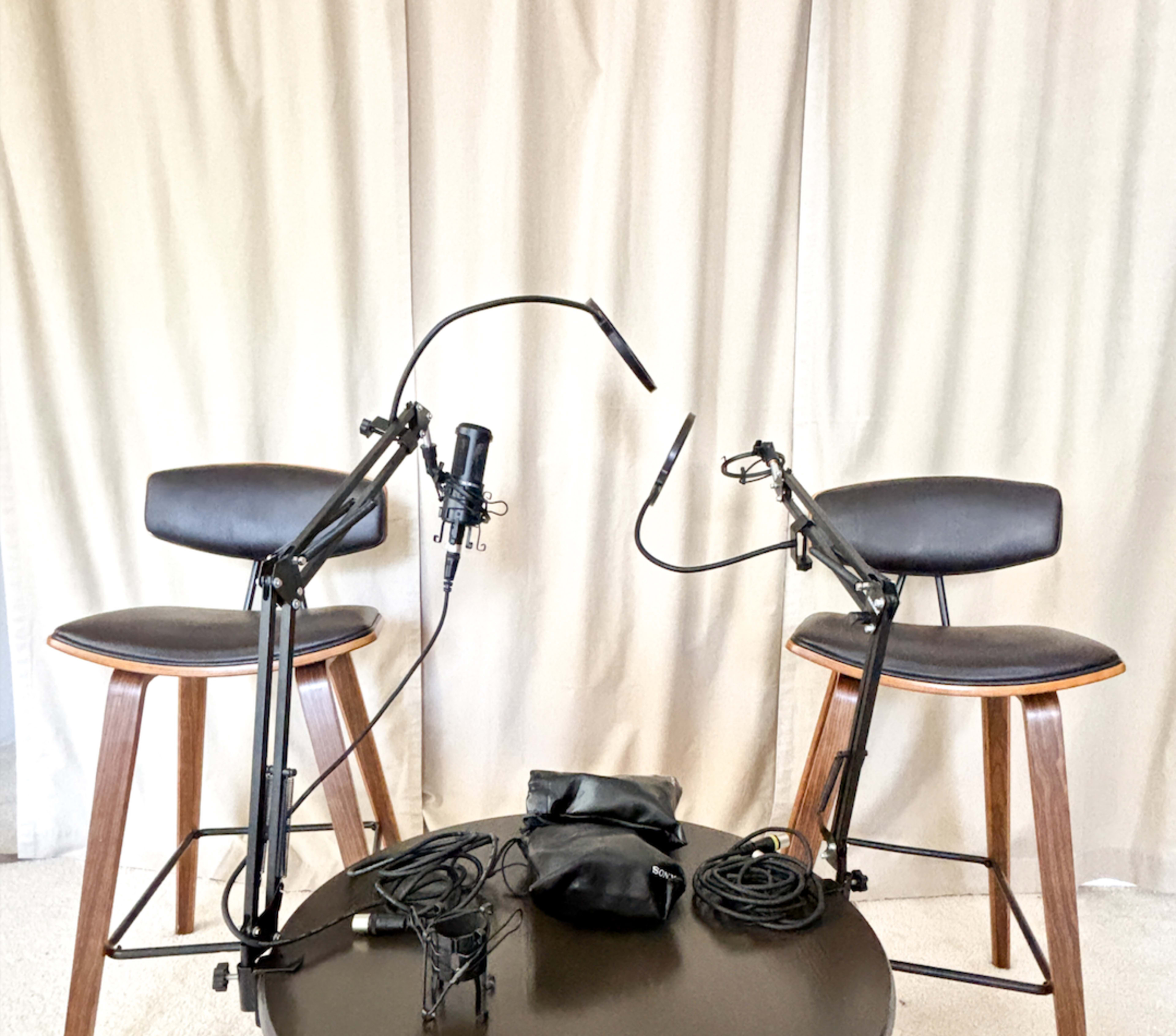 Two microphones mounted on adjustable arms are positioned in front of two wooden stools on a circular black table, with cables neatly placed on the table.