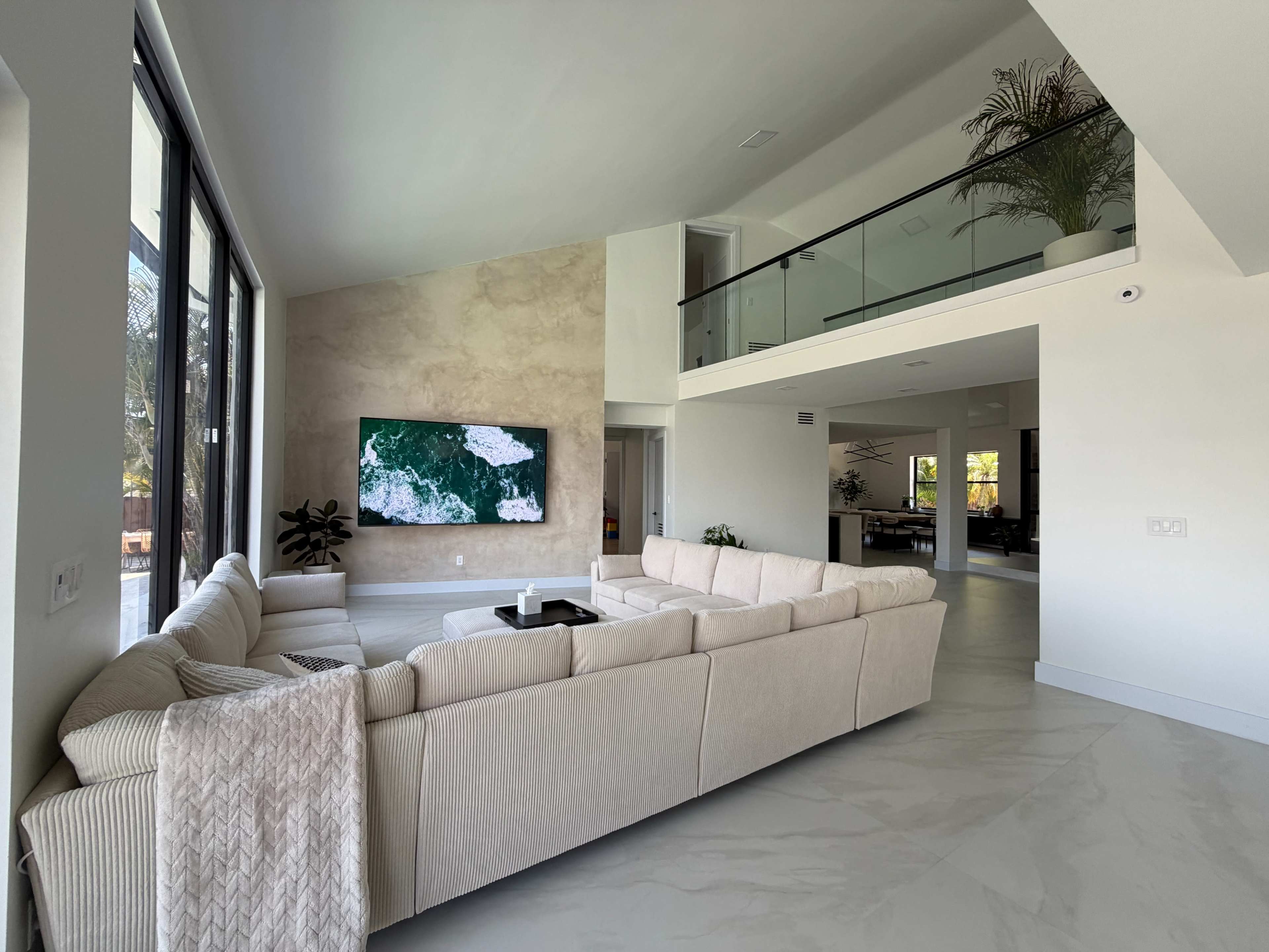 A spacious modern living room features a large sectional sofa, a mounted flat-screen TV on a textured wall, and an open view of an upper level overlooking the space.