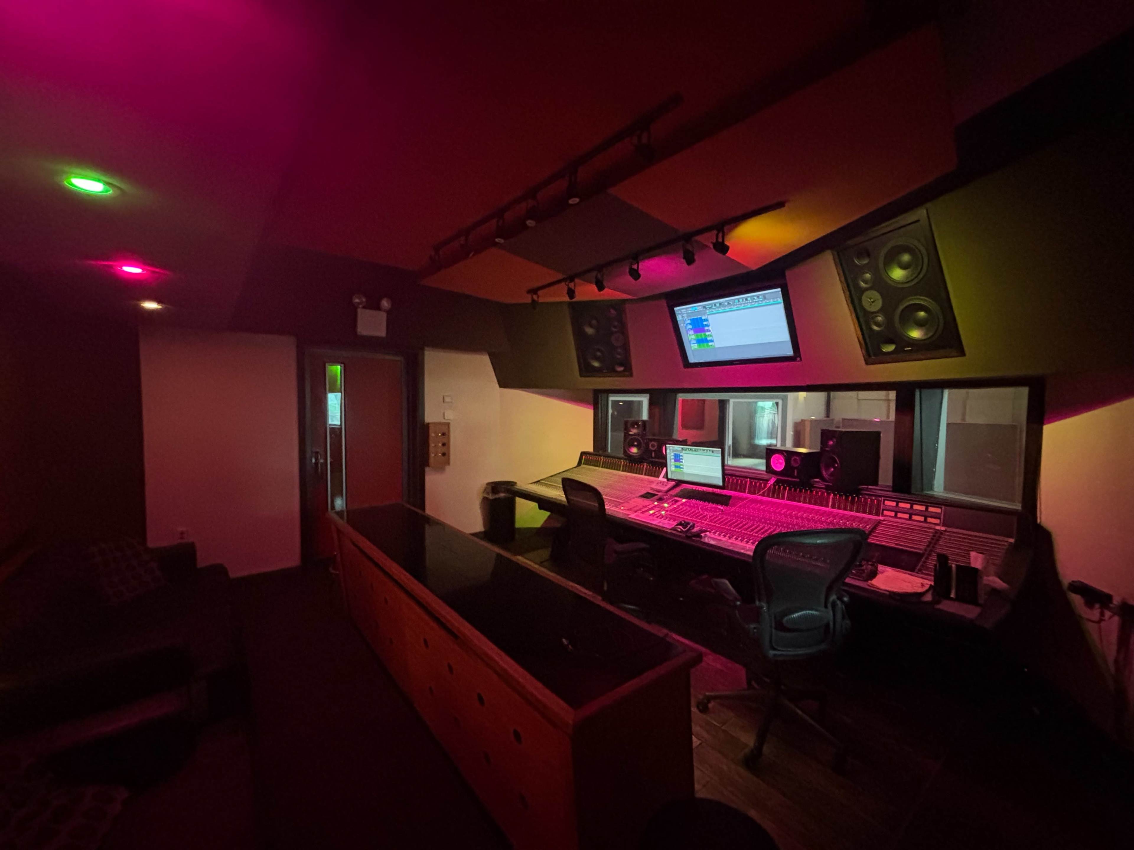 The image shows a sound recording studio with a central mixing console, speakers mounted on the walls, and colored lights illuminating the space.