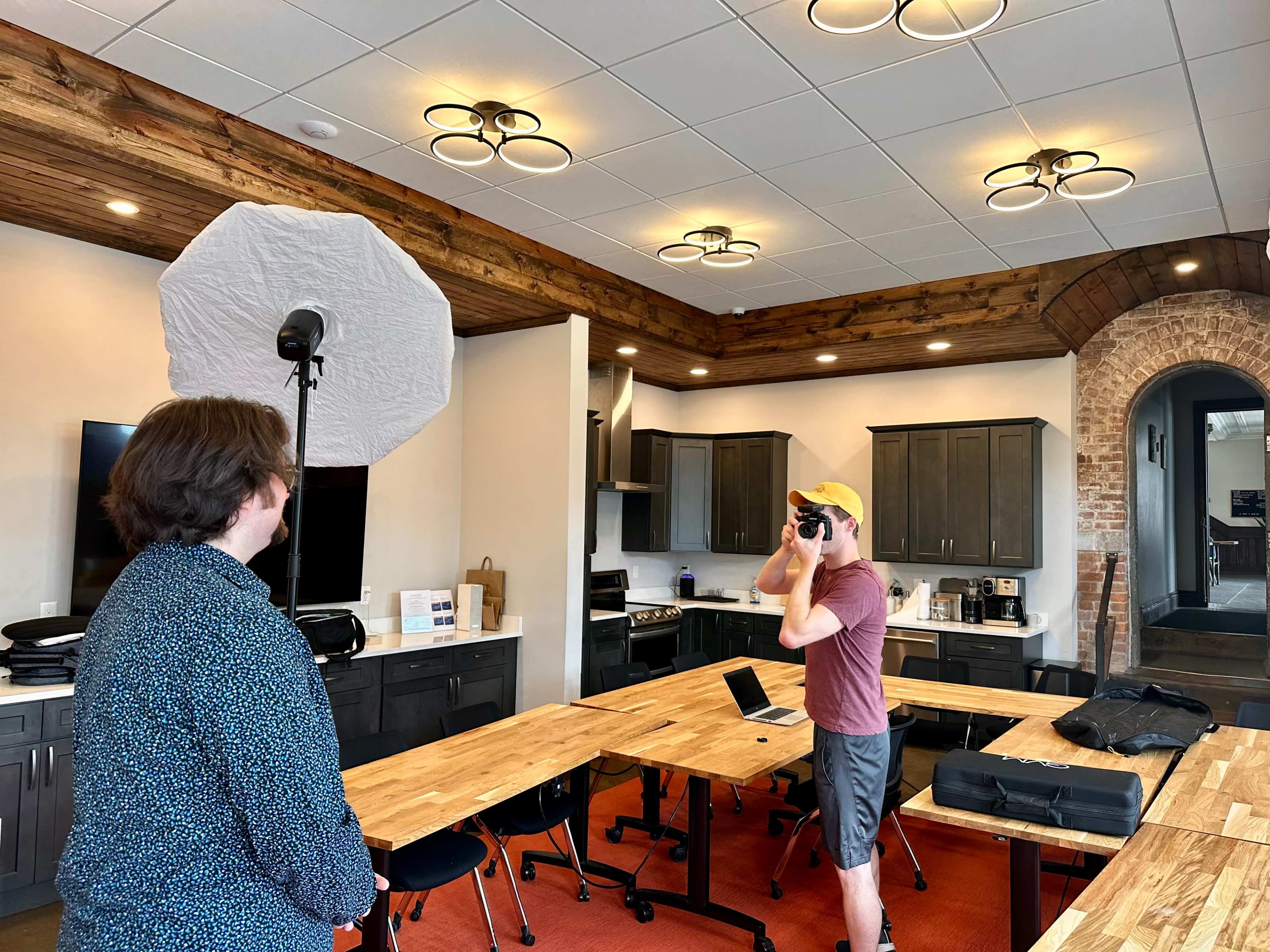 A man wearing a yellow cap takes photos of another man in a kitchen-like space equipped with wooden tables and modern appliances.
