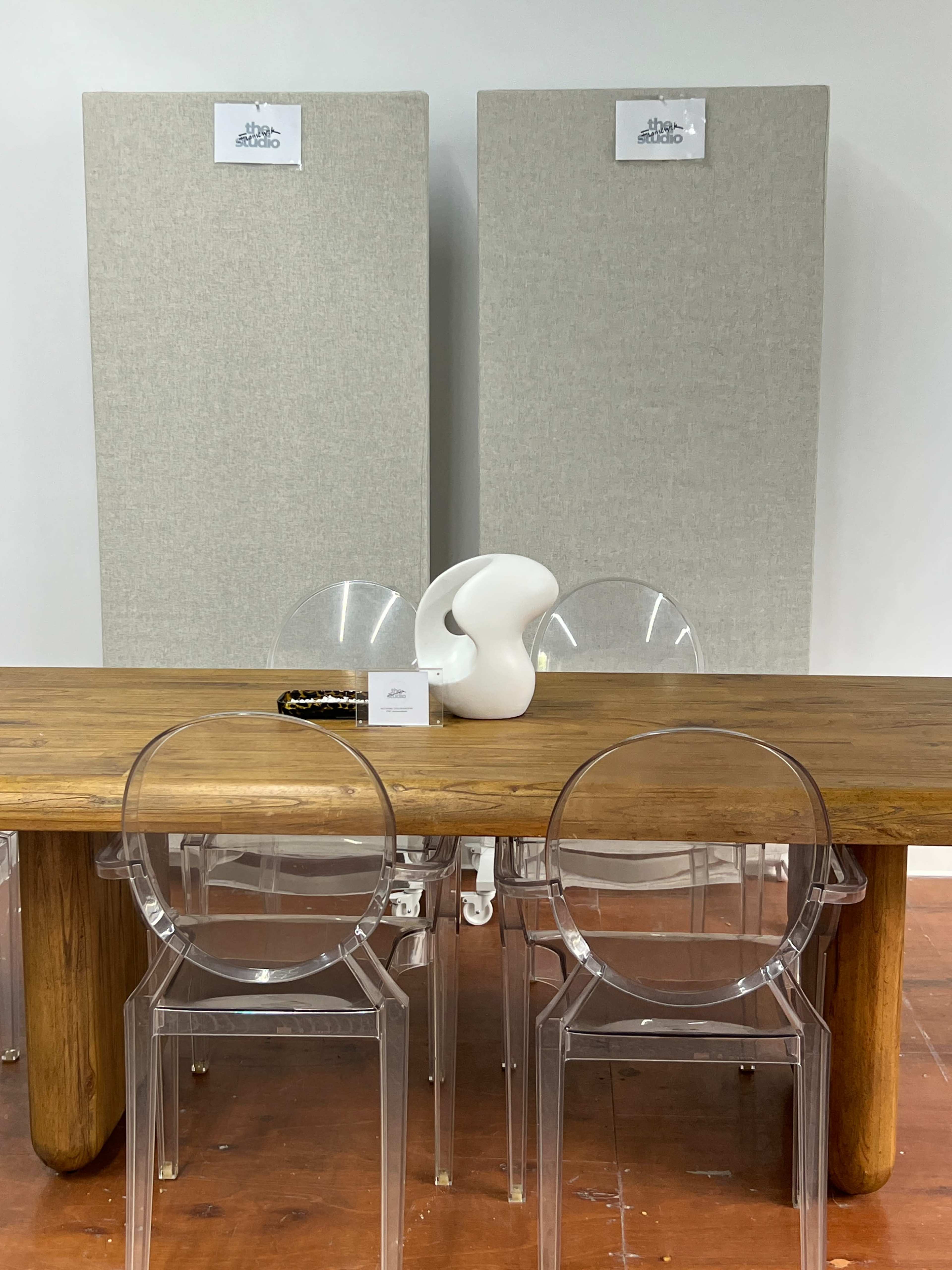 The image shows a wooden dining table with clear acrylic chairs in front of two tall, gray soundproof panels and a decorative white sculpture on the table.