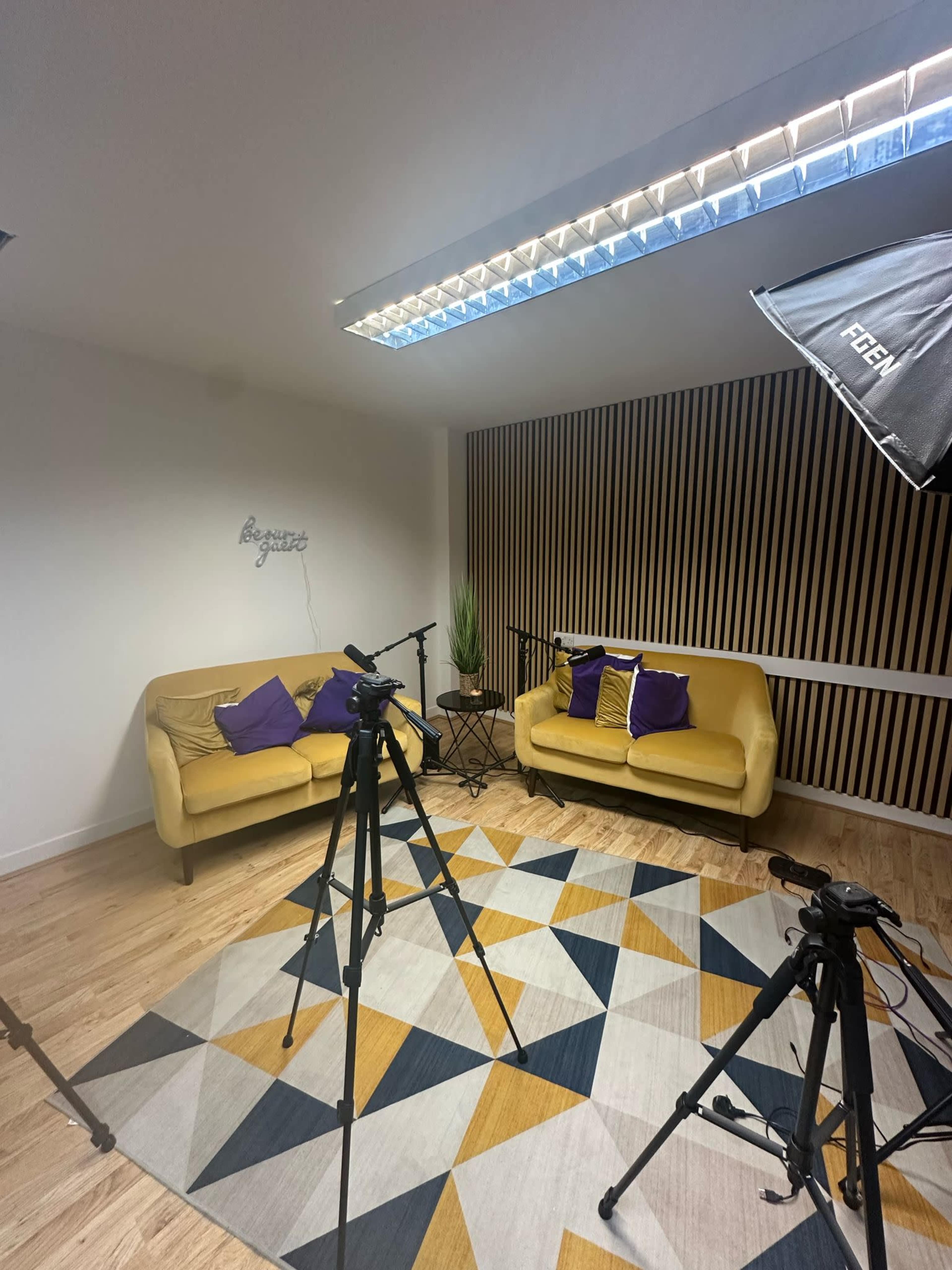 The image shows a studio setup with two yellow couches adorned with purple pillows, a patterned rug on the floor, and several camera tripods positioned around the seating area.