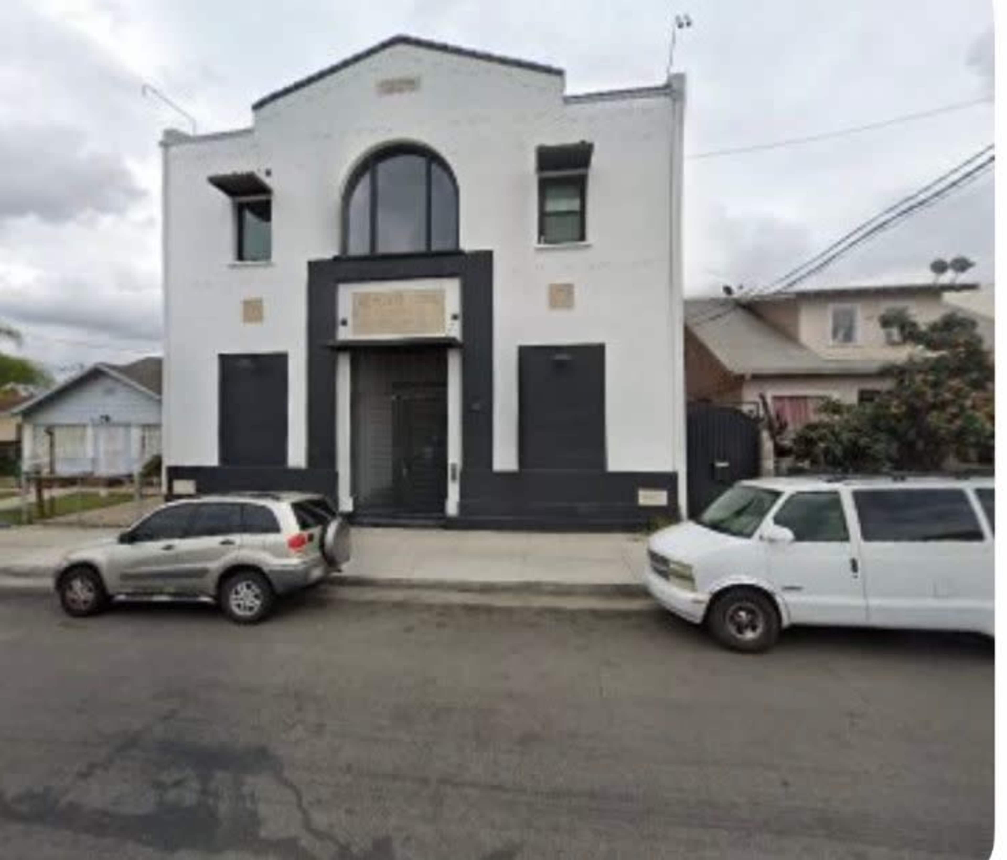 The image shows a two-story, white building with a distinctive arch above the entrance and two vehicles parked in front on a street.