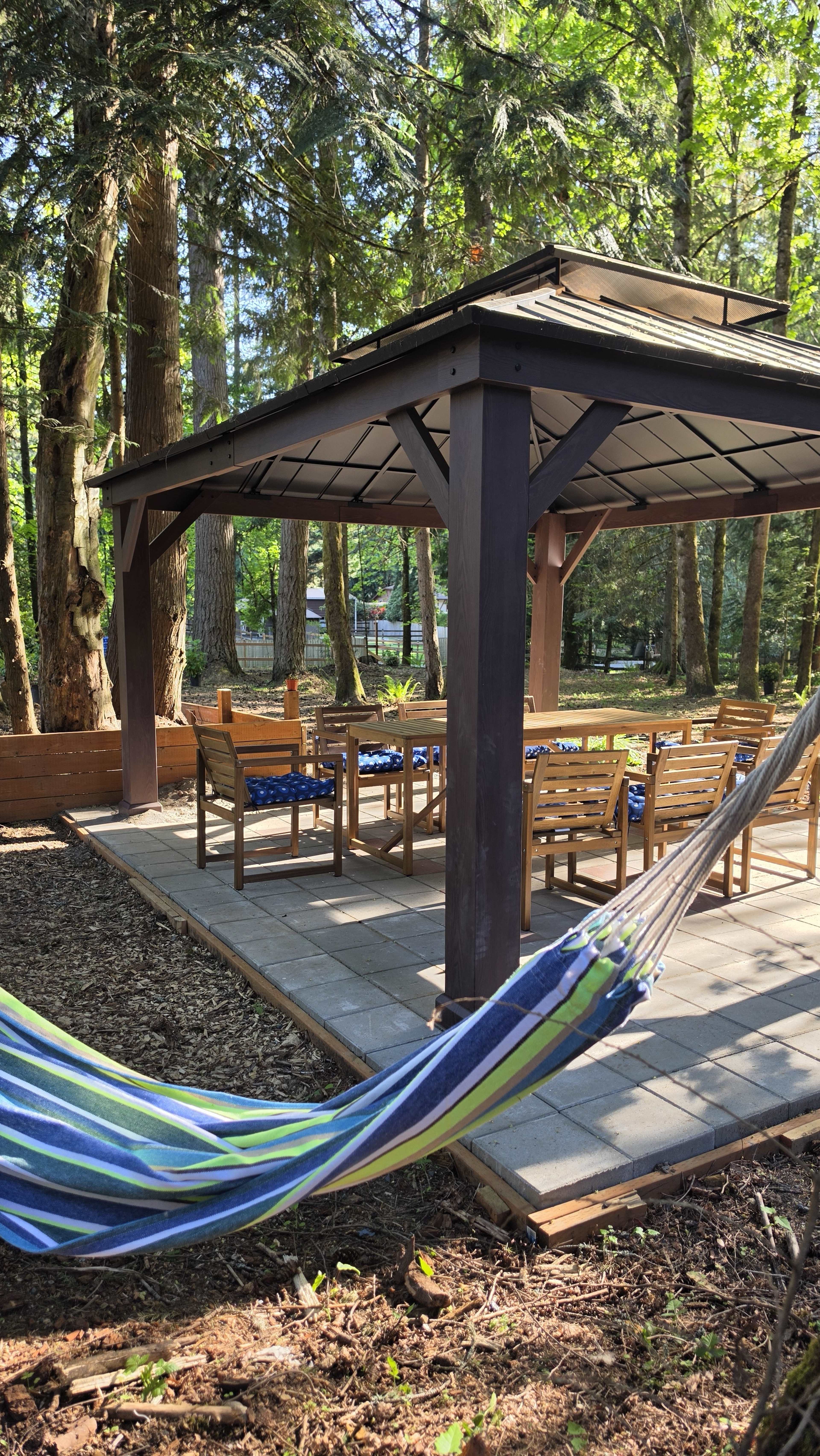 The image shows a gazebo with a table and chairs, surrounded by trees and featuring a hammock in the foreground.