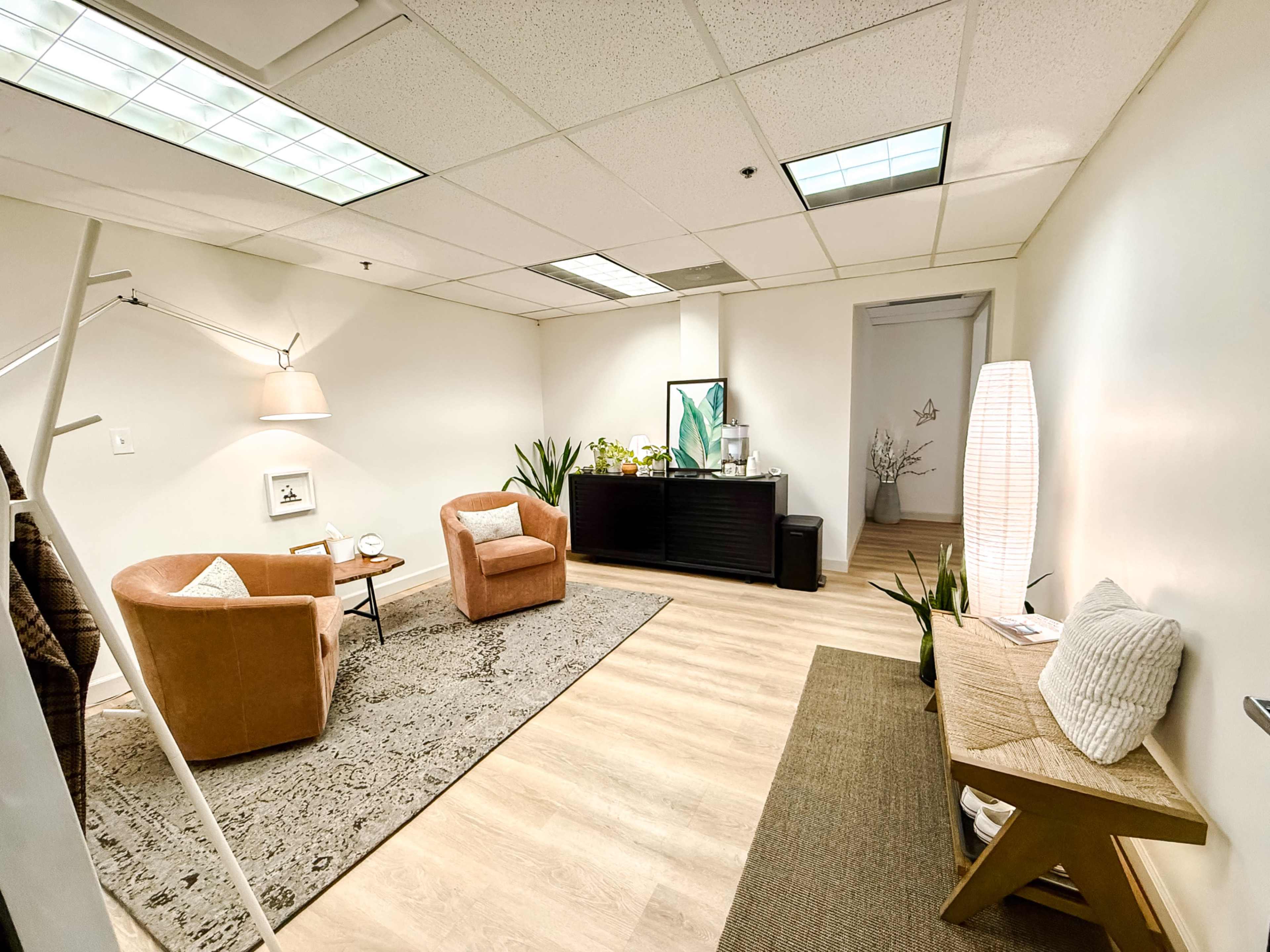 The image shows a bright, minimalistic office space featuring two tan chairs, a small side table, and a large mirror, along with decorative plants and a soft rug on the floor.