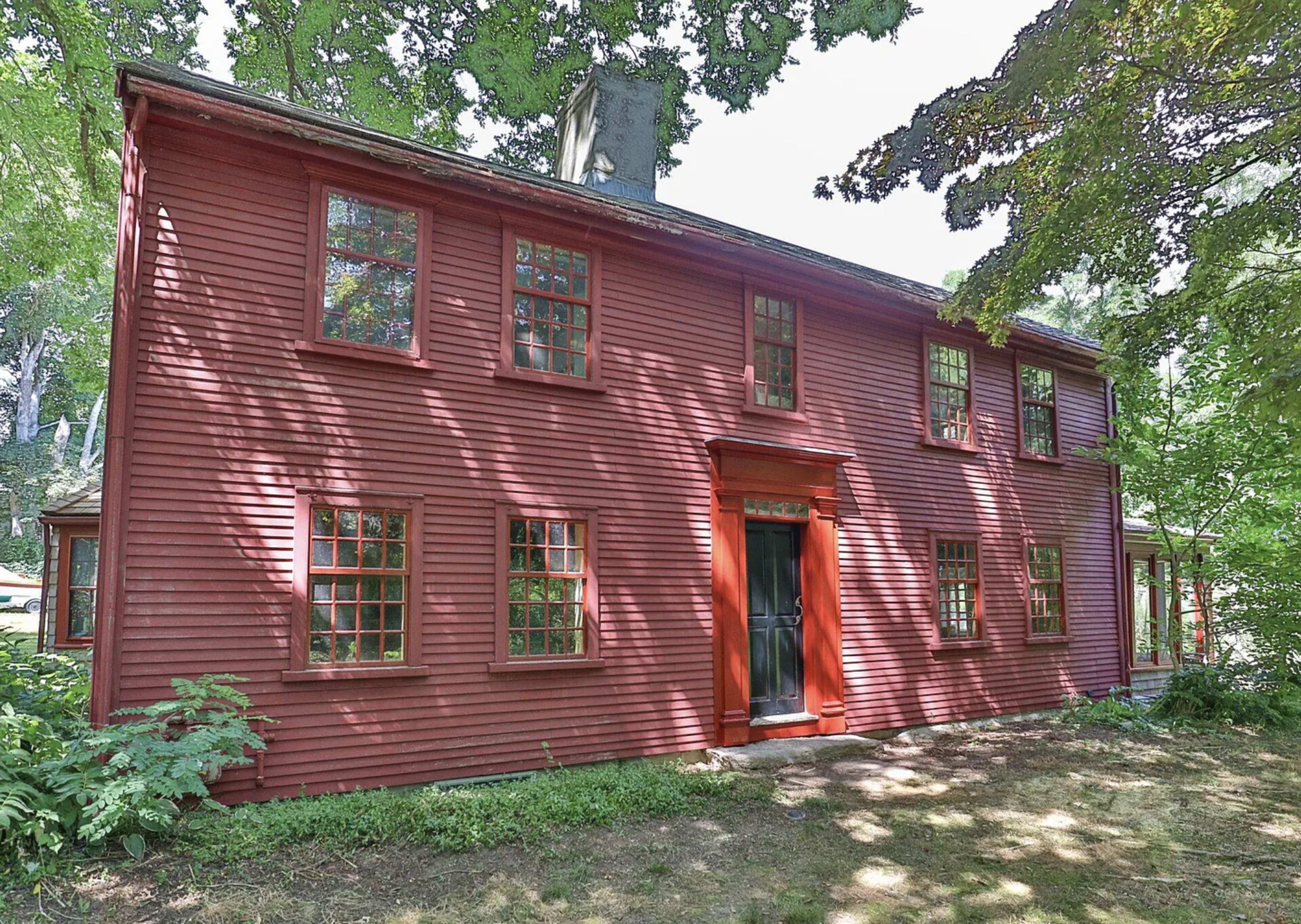 A two-story red wooden house with multiple windows and a central doorway, surrounded by greenery.