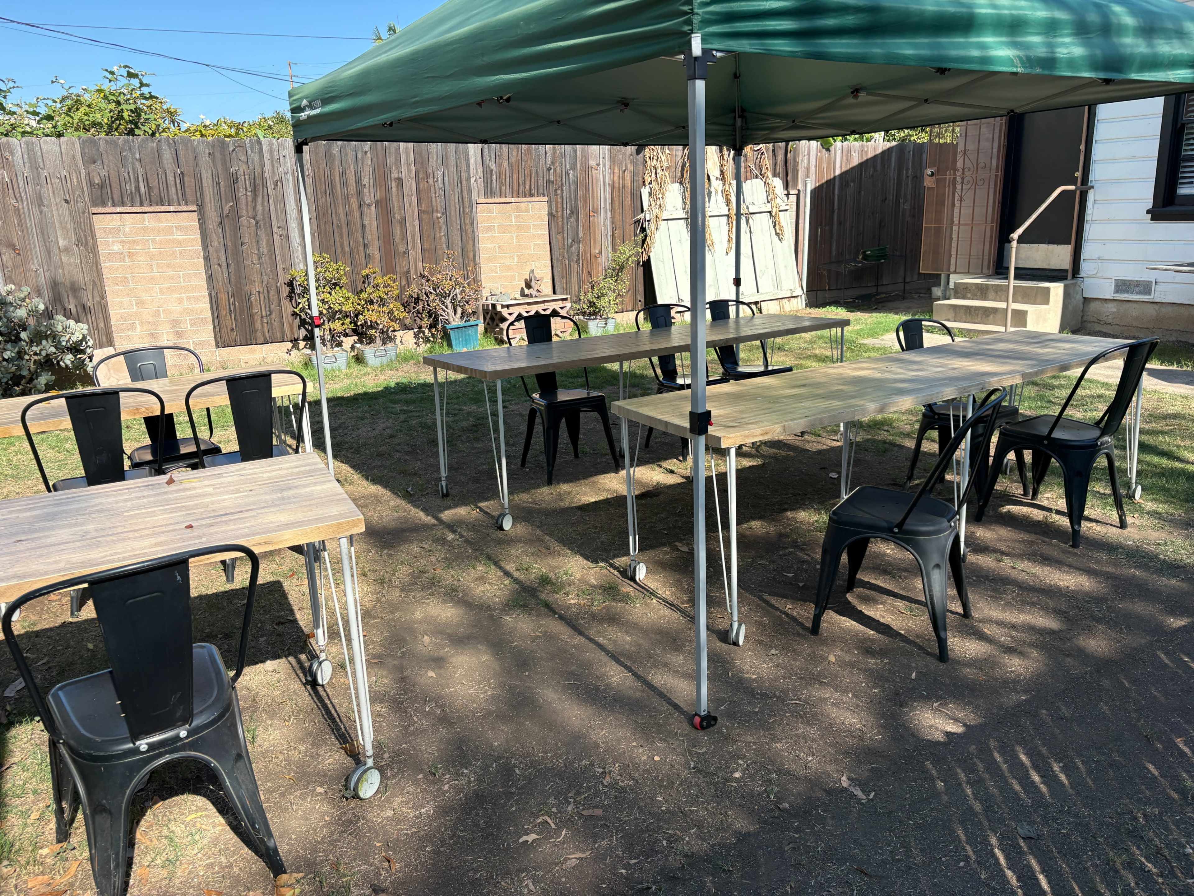 A green canopy covers several wooden tables and black chairs set up in a grassy outdoor area behind a house.