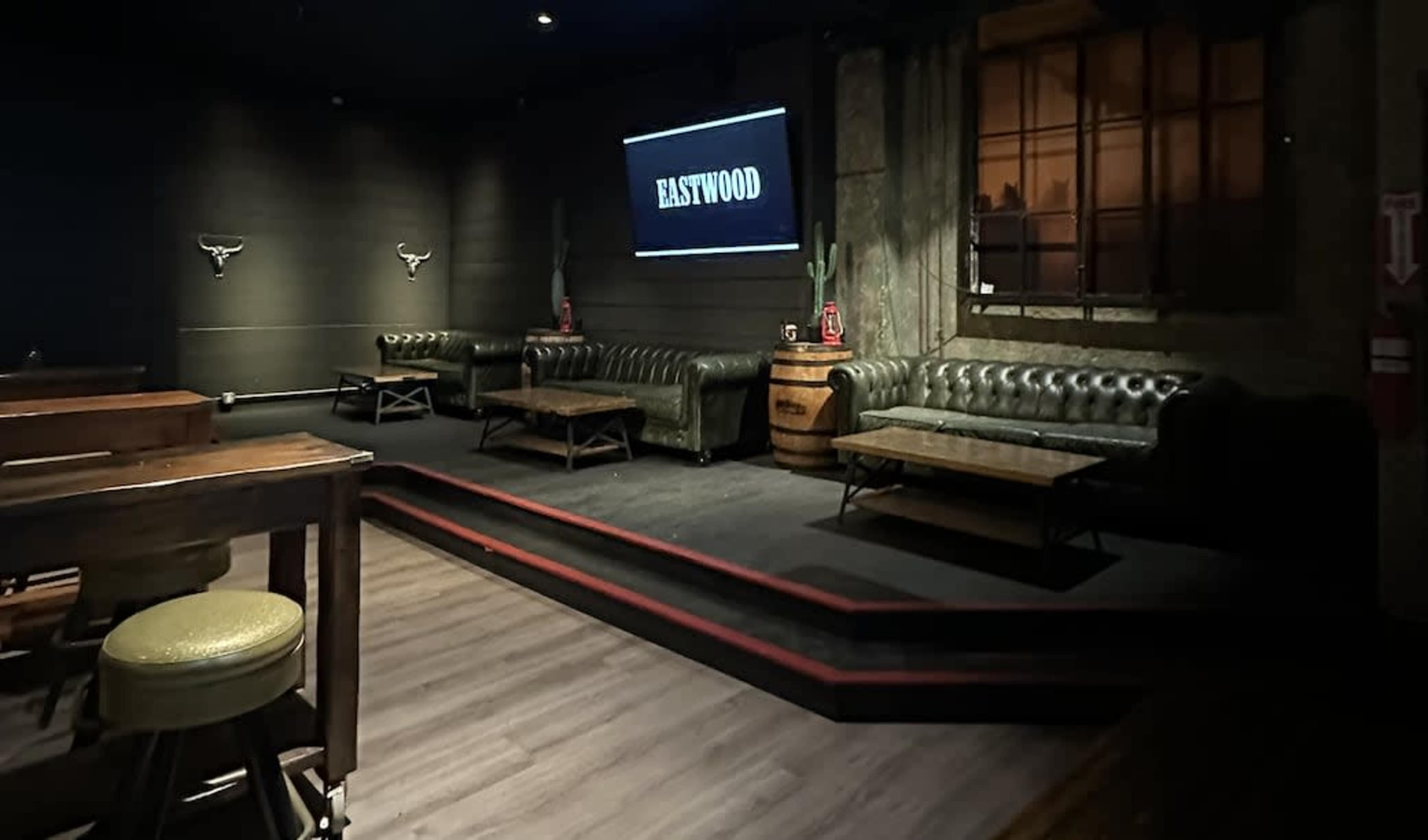 The image shows a dimly lit bar or lounge area with leather couches, a large screen displaying the word "EASTWOOD," and wooden tables, all set against a backdrop of brick walls and decorative cacti.