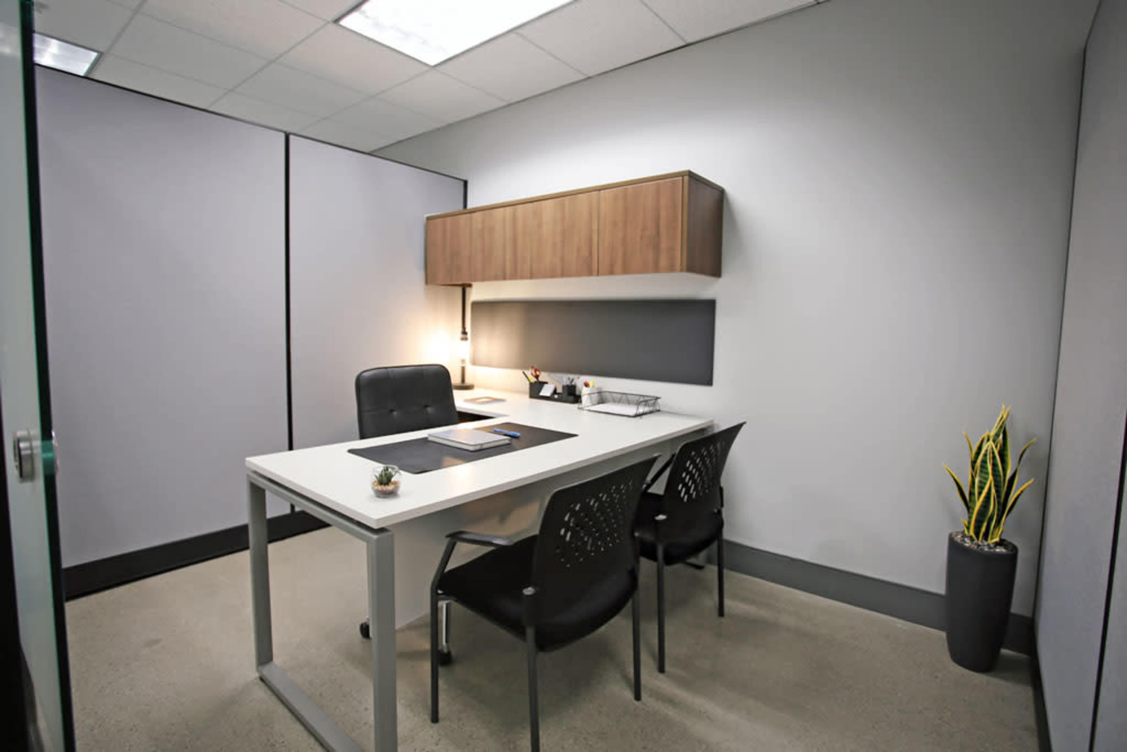 A minimalist office space featuring a white desk with two black chairs, a wall-mounted storage cabinet, and a potted plant.
