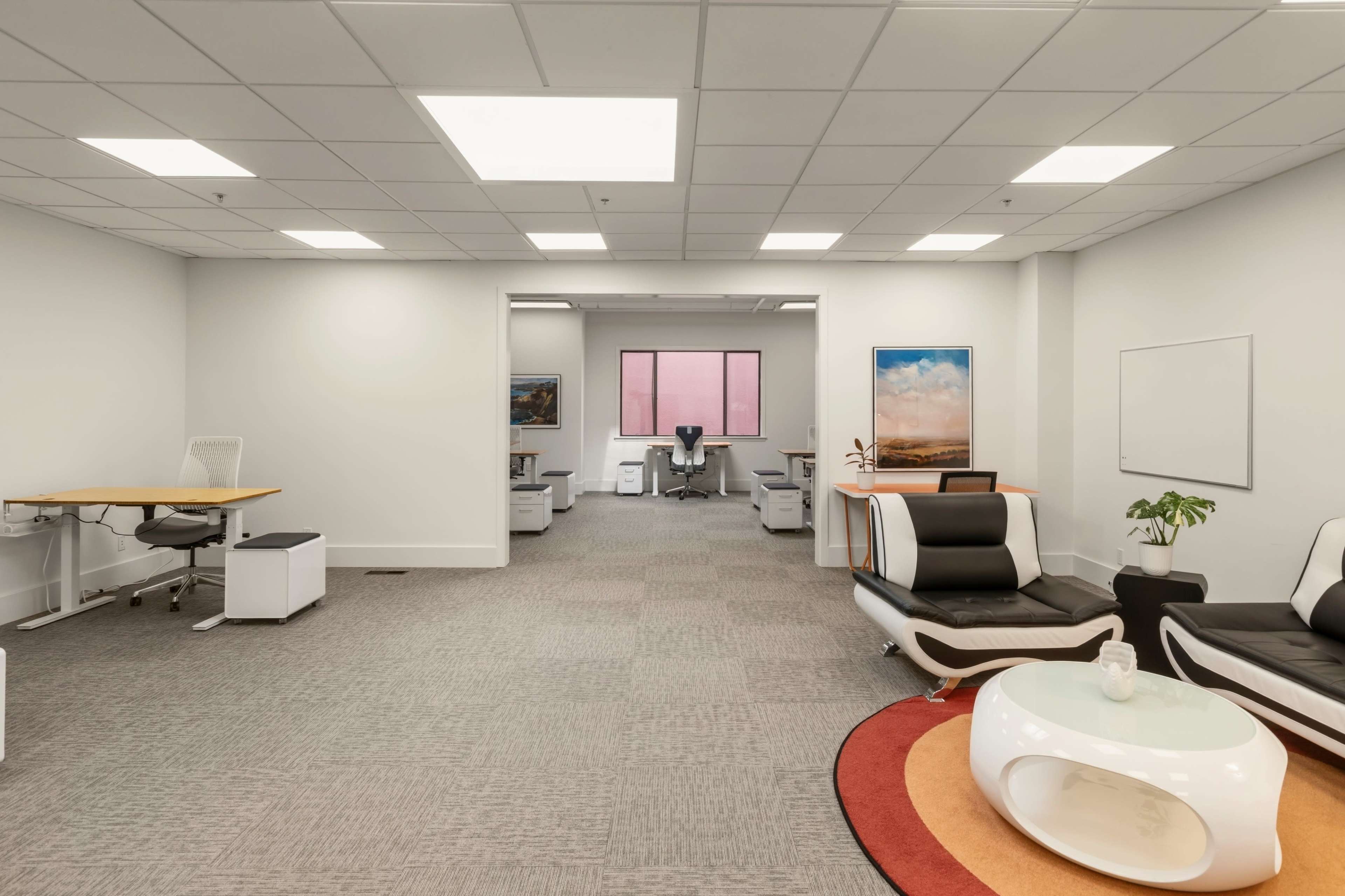 The image shows a modern office space featuring sleek furniture, including black and white seating, a circular coffee table, and several workstations in a well-lit environment.