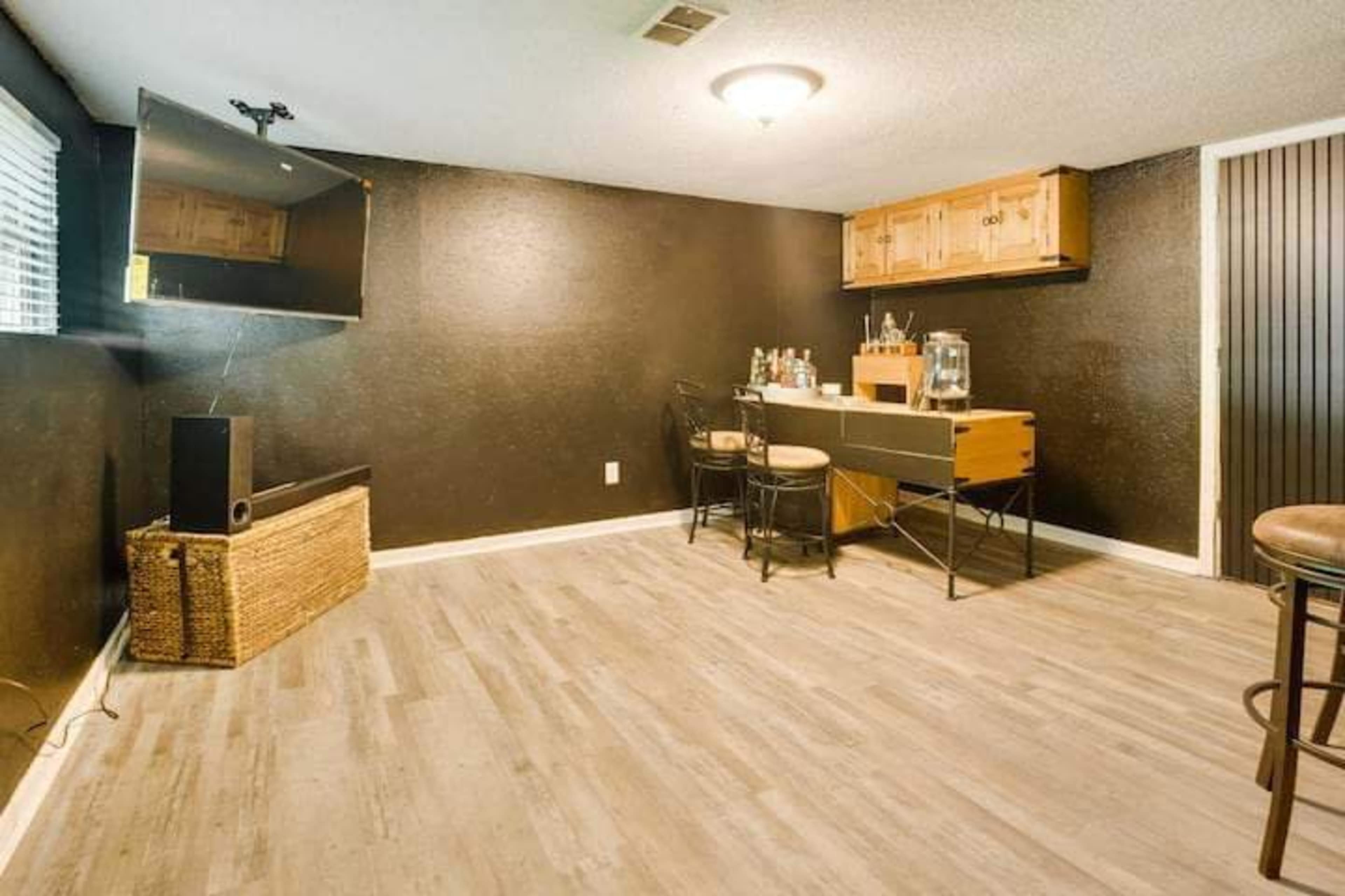 A dimly lit room with a bar setup featuring a table with a variety of bottles and two chairs, alongside a wall-mounted television and a woven storage basket.
