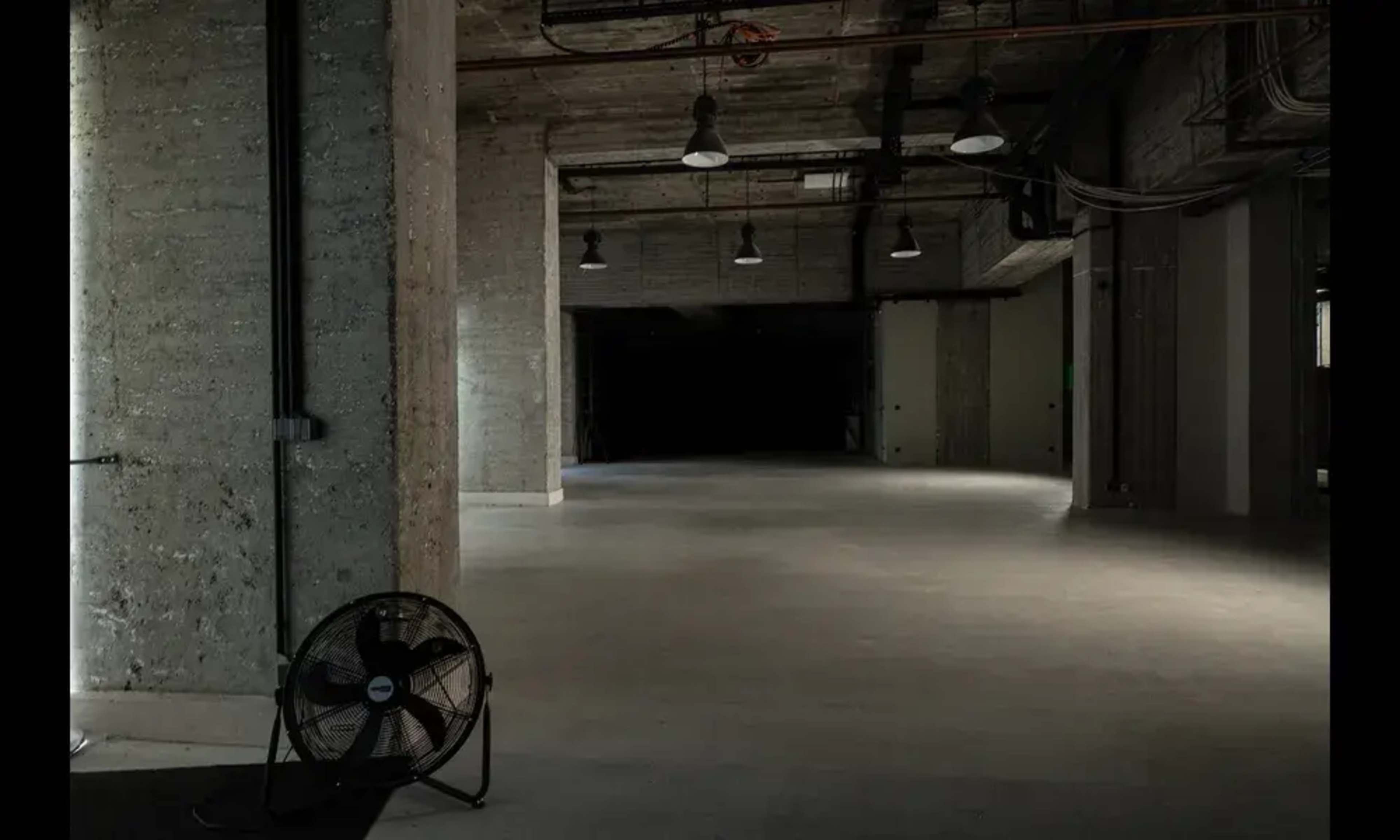 The image shows a large, empty interior space with concrete walls, low-light fixtures, and a standing industrial fan in the foreground.