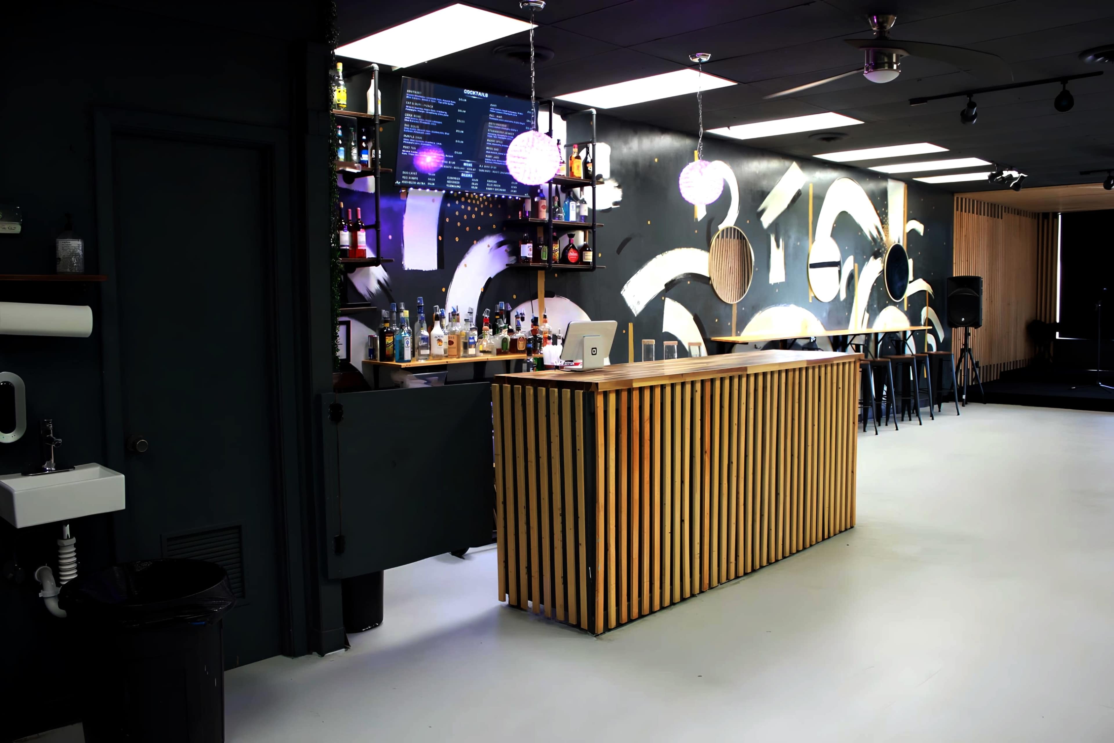 A modern bar features a wooden counter, illuminated shelves of liquor, and a minimalist design with geometric wall graphics.