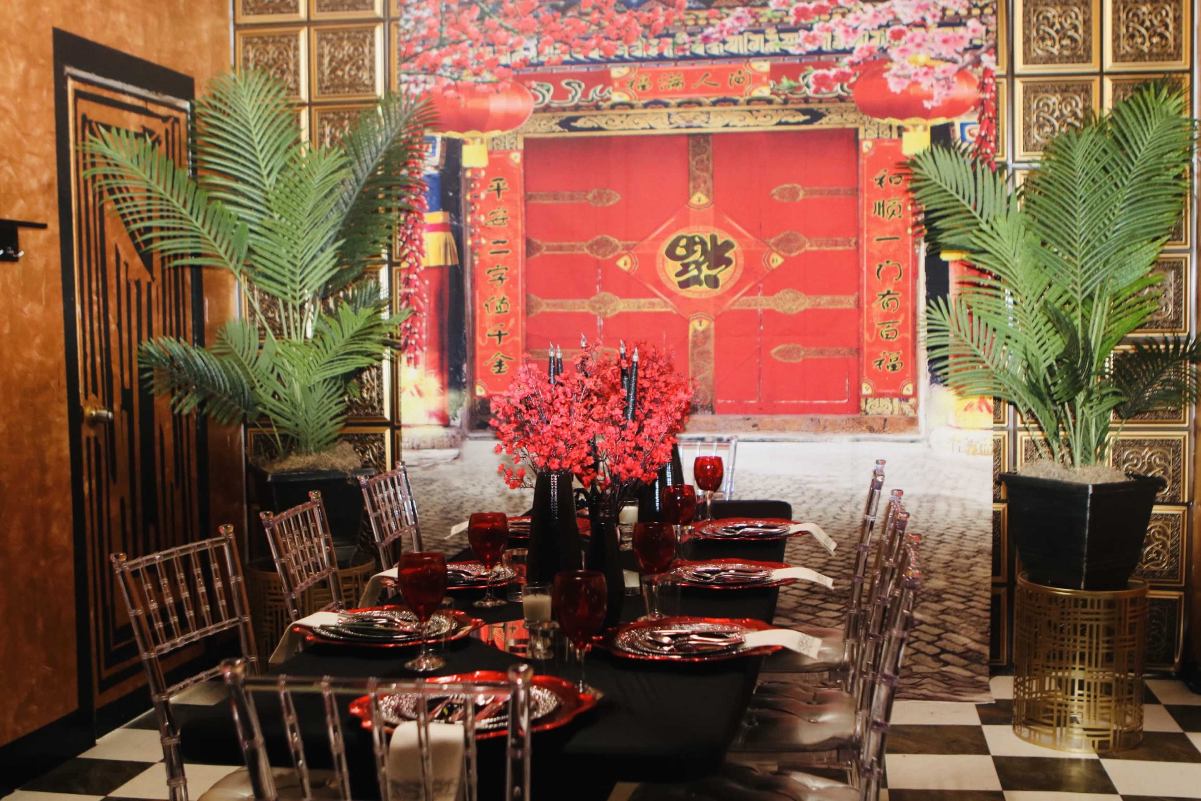 A dining table is set with black and red dinnerware in front of a vibrant mural depicting a traditional red door adorned with flowers and lanterns.