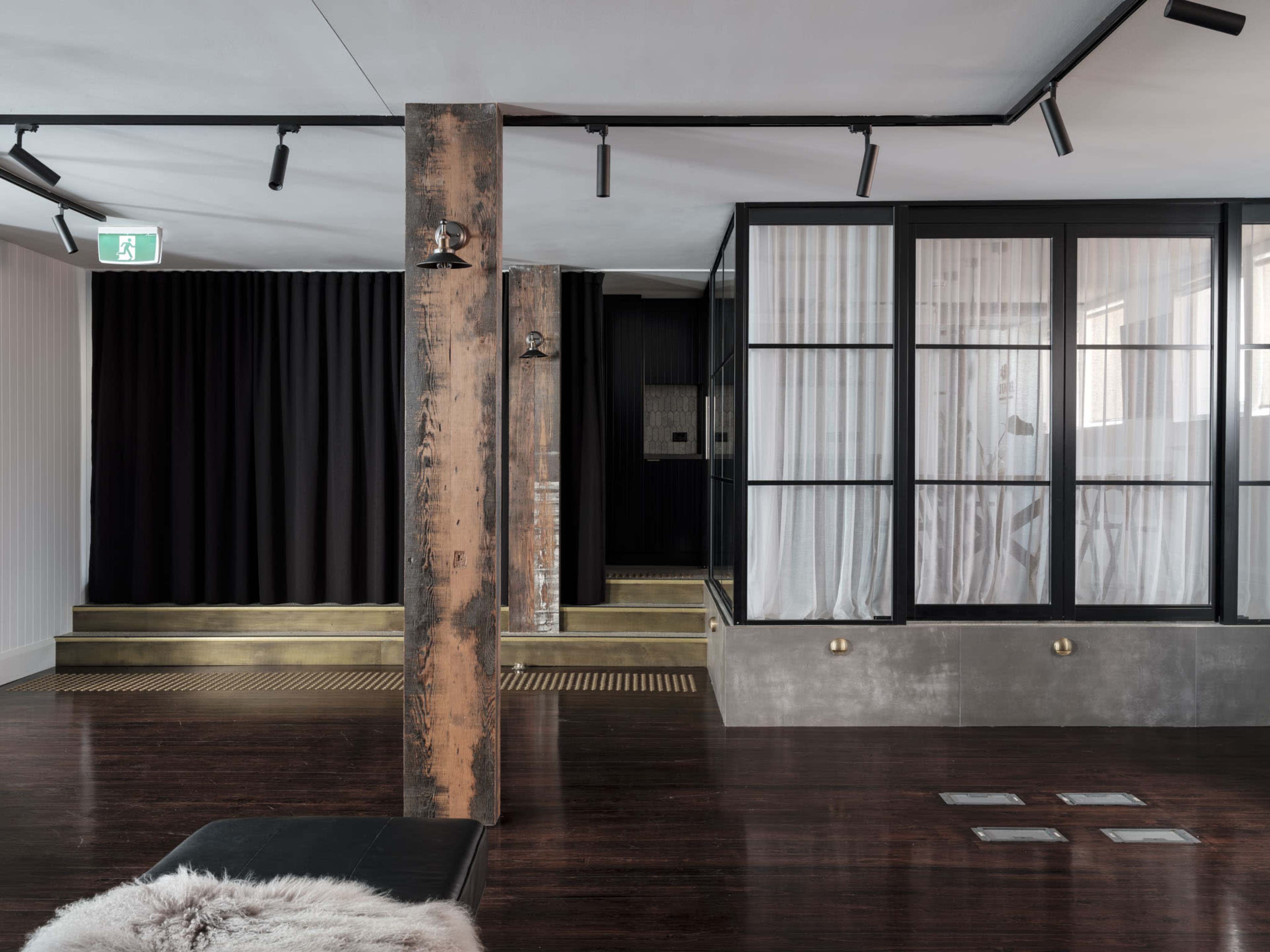 The image shows a modern interior space featuring a wooden floor, a prominent metal column, black curtains, and glass partitions that create separate areas.